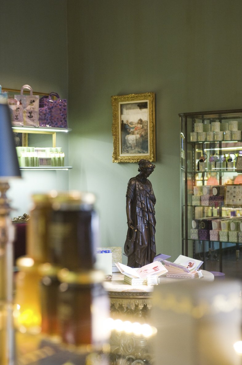 Une boutique avec des étagères remplies de produits, une statue en bois d'une femme, une peinture encadrée gold, et des papiers sur une table.