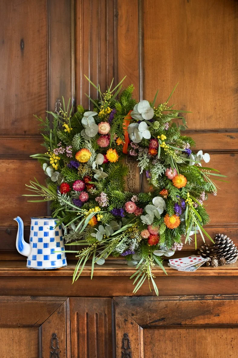Un mur en bois avec un guirlande de fleurs colorées accrochée au centre, un petit pichet en céramique à carreaux bleus et blancs, une paire de pommes de pin, et un jeu de cartes sur une étagère en bois.
