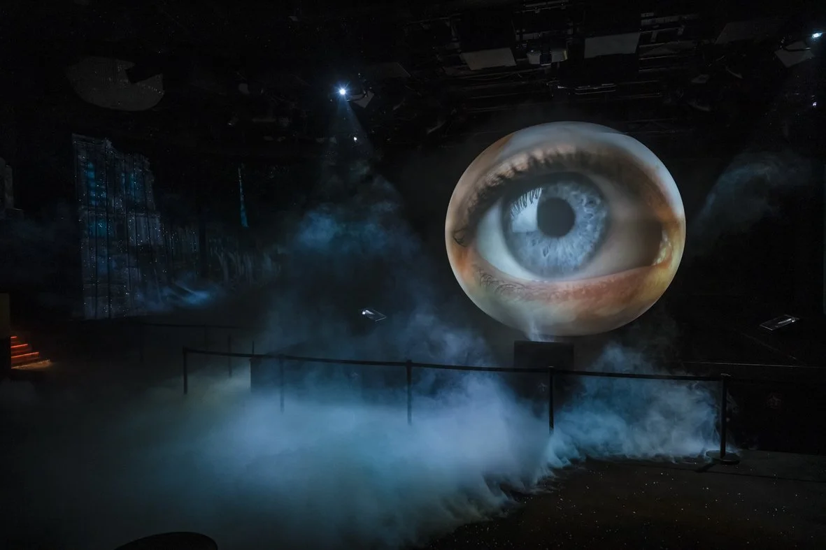 Une scène sombre avec un grand œil projeté en arrière-plan, entourée de brouillard et d'illuminations bleues, donnant l'impression d'une atmosphère mystérieuse ou dramatique.