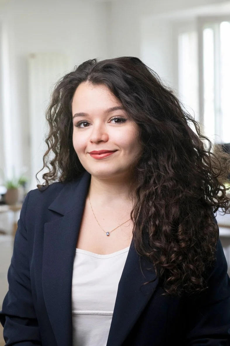 Portrait d'une femme avec des cheveux bouclés, portant une veste bleue et un collier en or, dans un environnement intérieur lumineux.