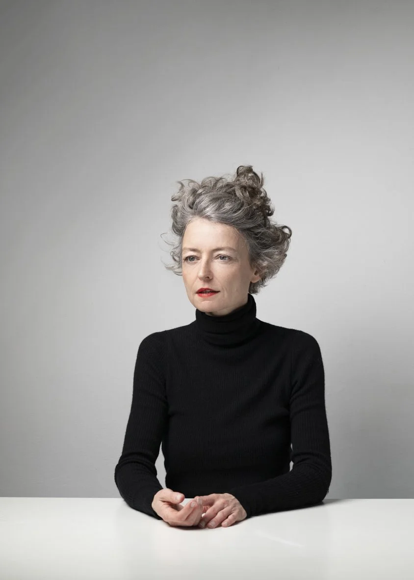 Une femme âgée avec des cheveux gris bouclés, portant un turtleneck noir, assise à une table blanche, regardant légèrement sur le côté, avec un fond gris clair.