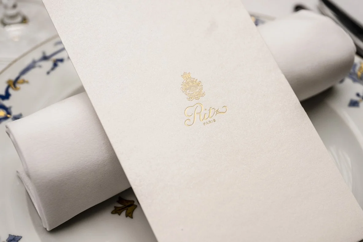 Une boîte blanche avec un logo doré de la marque 'Ritz Paris' placée sur une table avec une nappe colorée en arrière-plan.