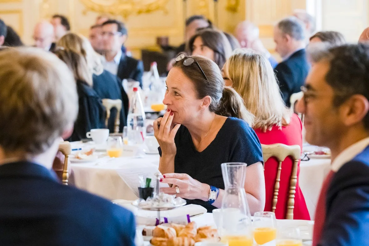 Groupe de personnes assises autour d'une table lors d'un déjeuner ou d'une réunion, dans une salle ornée de bois, en train de parler et d'écouter, avec des boissons et de la nourriture sur la table.