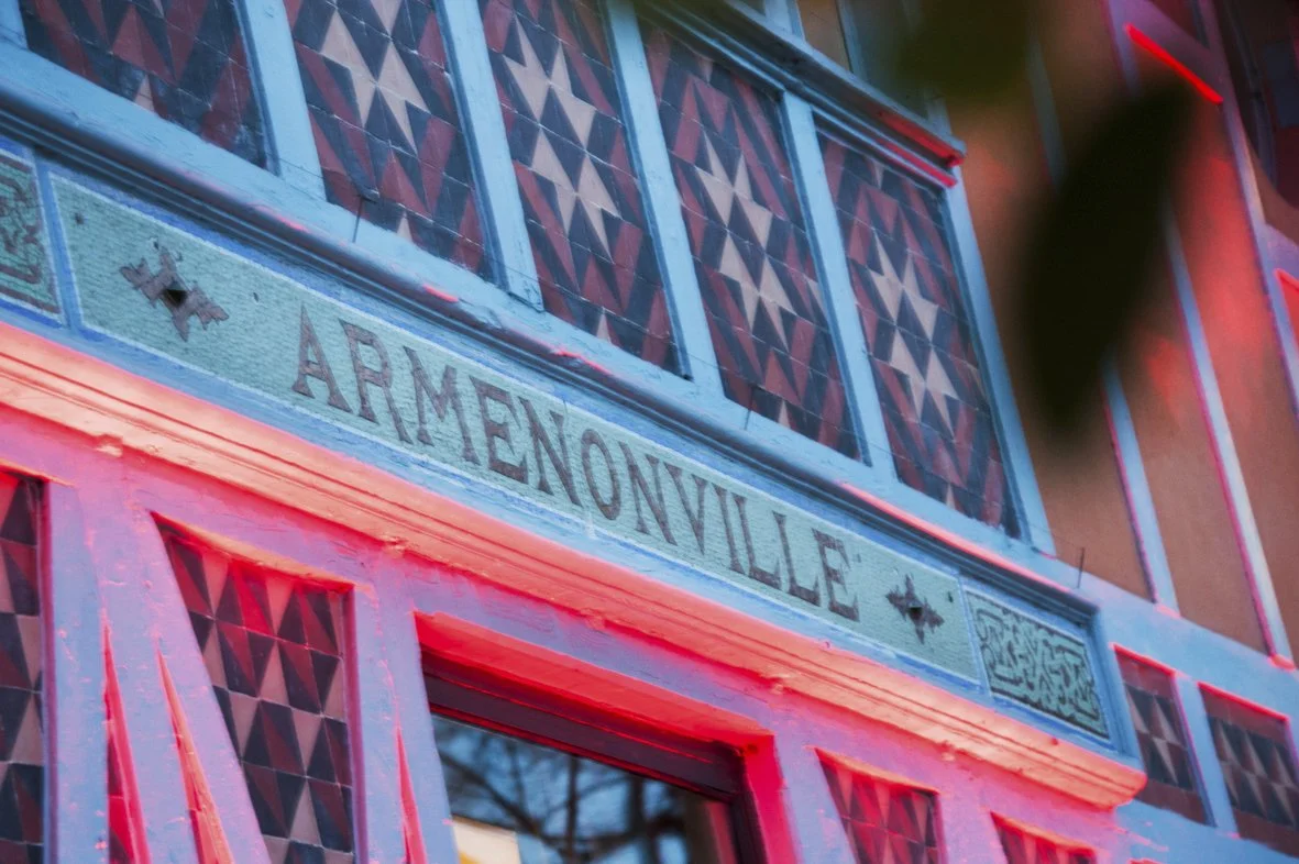 Façade colorée avec le nom Arménoville écrit sur un panneau, avec des fenêtres aux motifs géométriques en arrière-plan, éclairée par des lumières rouges et bleues.