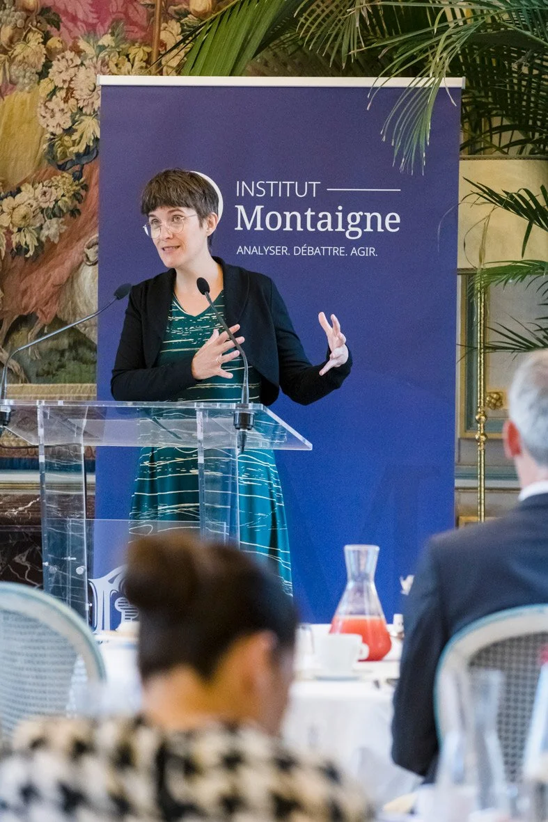 Une femme parle lors d'une conférence à l'Institut Montaigne, dans une salle ornée de décorations florales et de plantes, avec une bannière bleue à l'arrière
