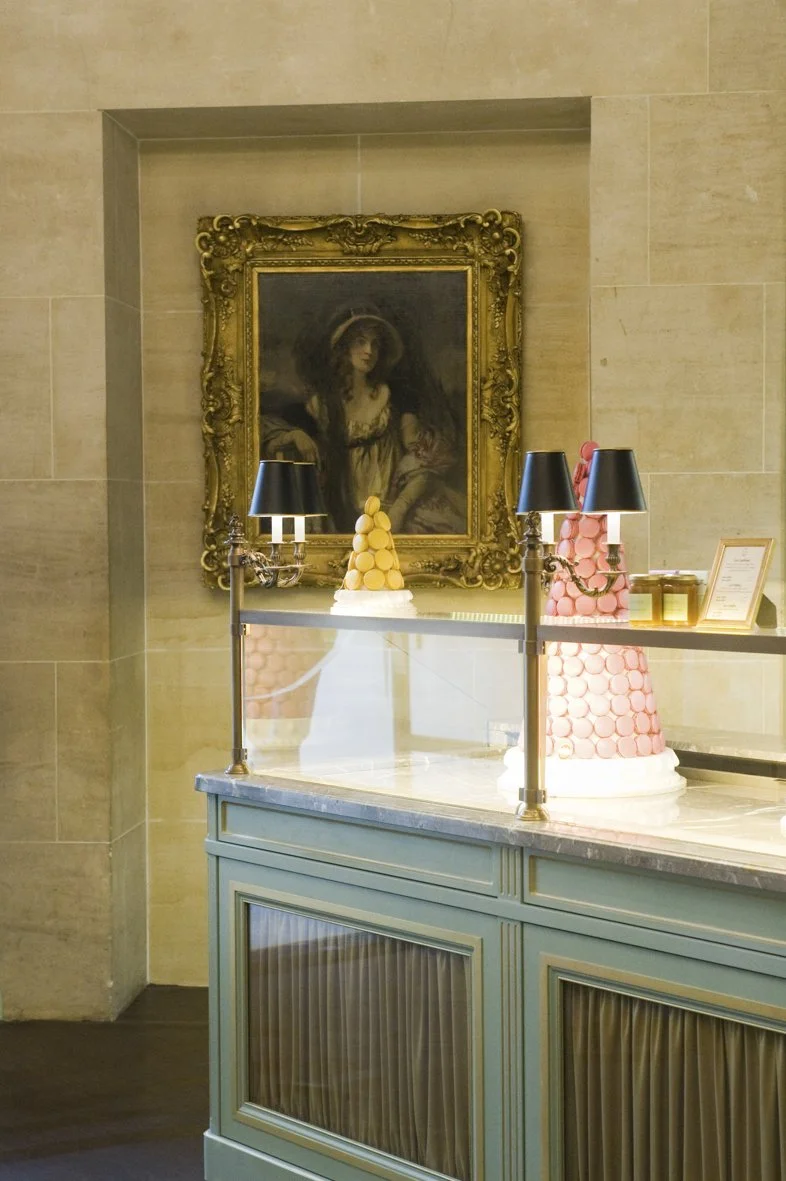 Deux desserts en forme de pyramide, décorés de macarons colorés, sont placés sur un comptoir en marbre dans une décoration intérieure élégante. Un tableau ancien orné d'un cadre doré est accroché au mur derrière le comptoir.