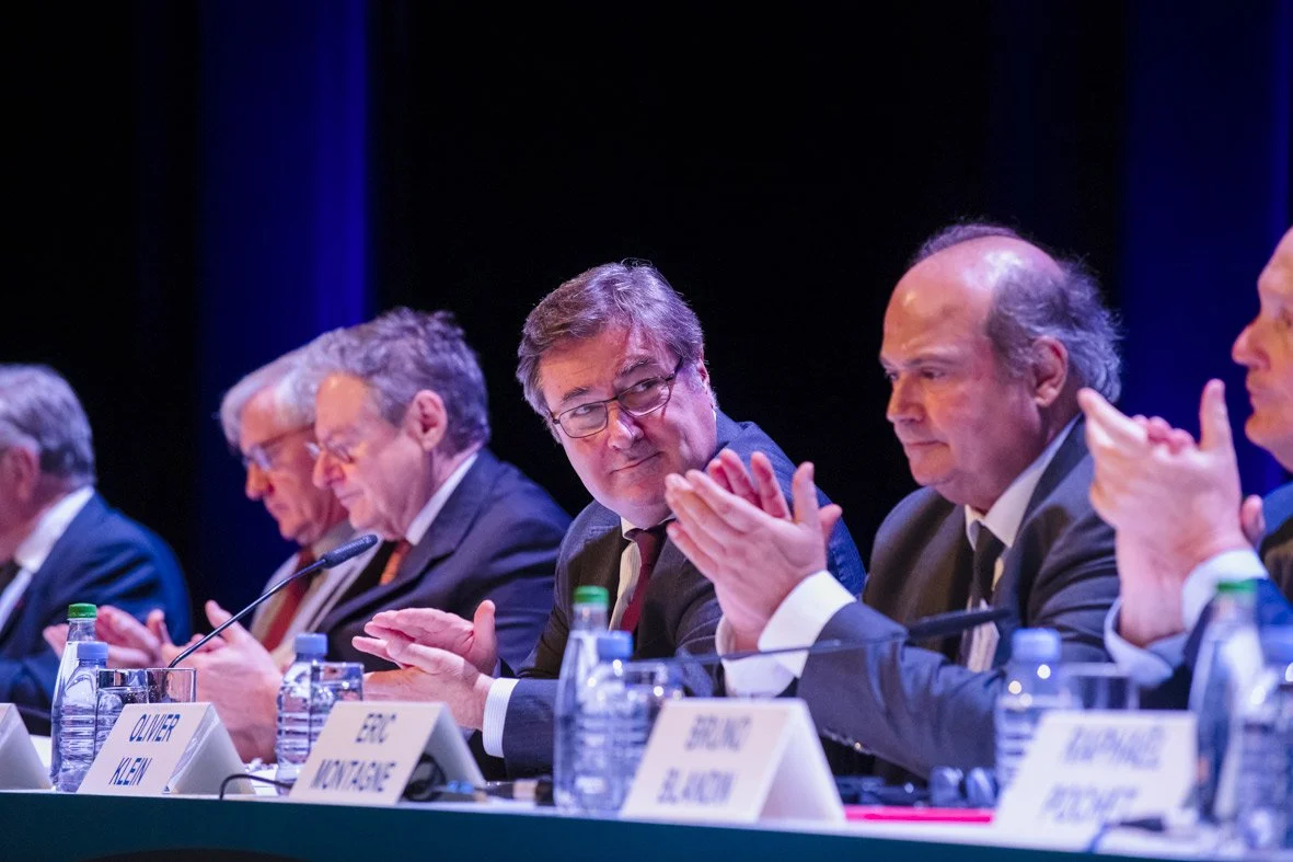 Groupe de personnes assises à une table lors d'une conférence, en train de discuter et de prendre des notes, avec des bouteilles d'eau devant eux.