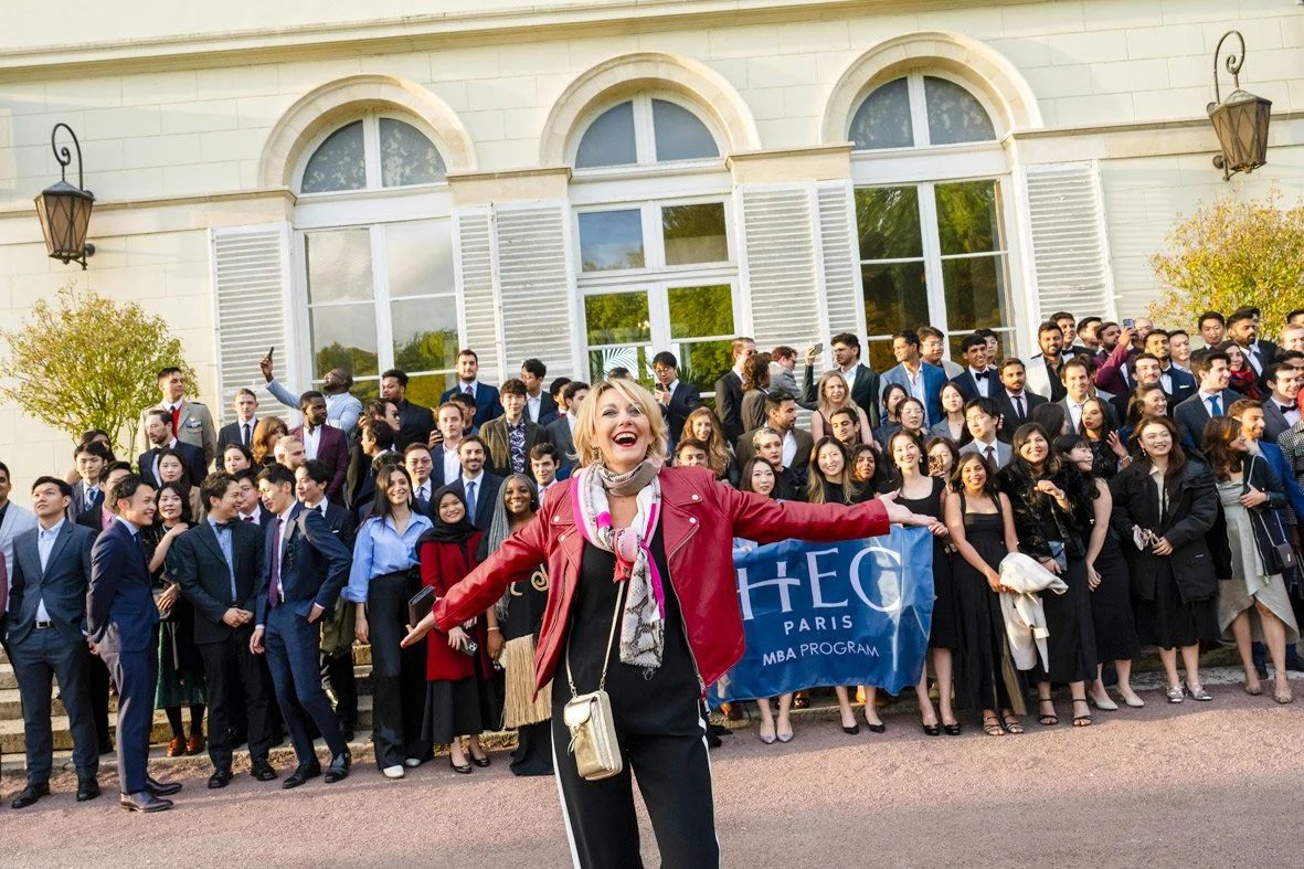 Un groupe de jeunes étudiants posant devant un bâtiment avec une femme souriante au premier plan, tenant un drapeau de l'ESCP Paris. L'ambiance semble joyeuse et festive.