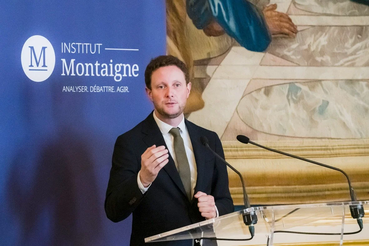 Un homme en costume noir parle devant un micro à une conférence organisée par l'Institut Montaigne, avec un tableau ou un mur en arrière-plan.
