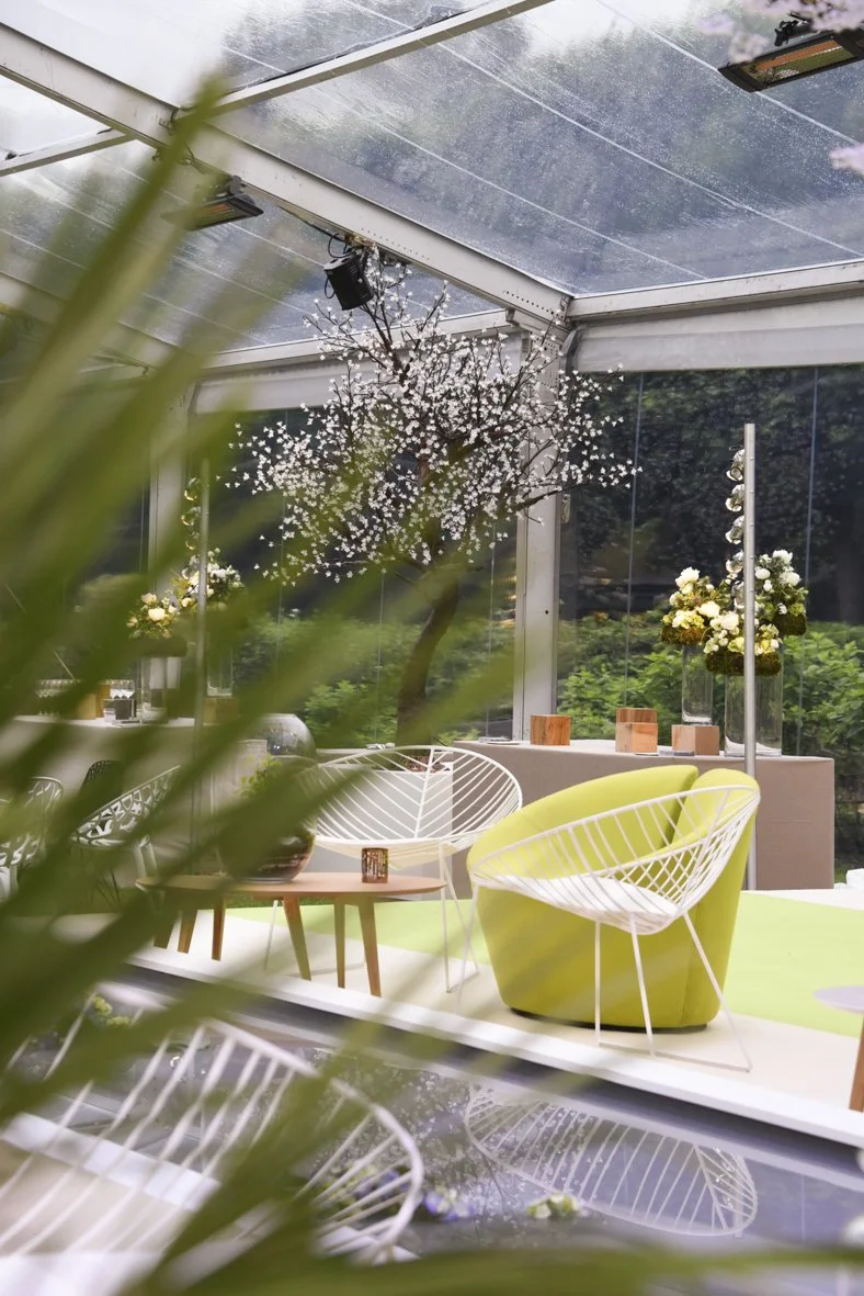 Intérieur moderne avec fauteuils en plastique blanc et vert, table en bois, fleurs dans des vases, arbre en fleurs derrière.