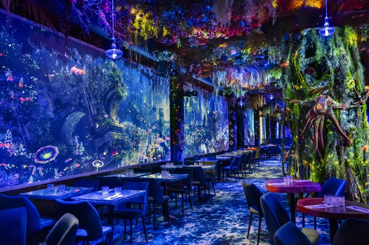 Restaurant intérieur décoré de plantes et de lumières colorées, avec des tables, des chaises et une grande fresque digitale représentant une forêt enchantée.