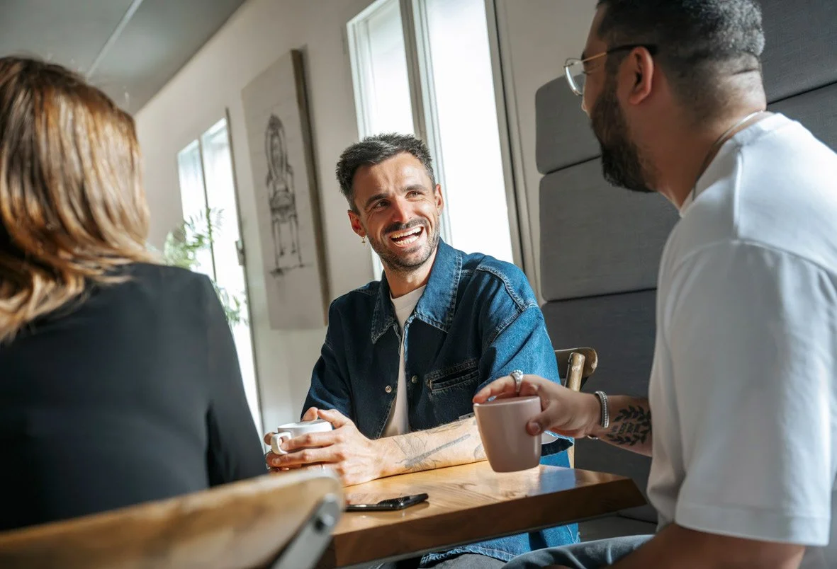 Trois personnes discutent et sourient dans un café, deux hommes et une femme, assis autour d'une table en bois, en train de boire du café.