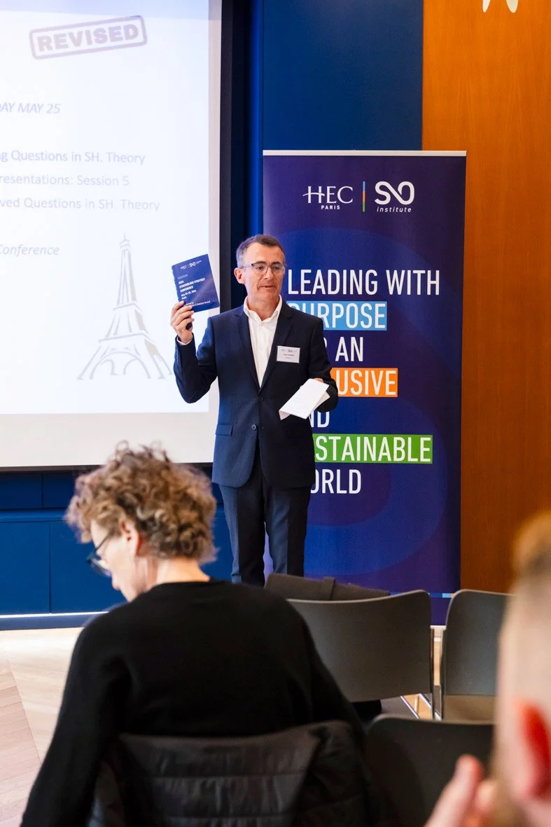 Un homme en costume parle lors d'une conférence, tenant une brochure dans sa main, avec un pupitre à côté et un drapeau en arrière-plan avec le texte 'Leading with Purpose' et des mots 'Exclusifs' et 'Durables'.