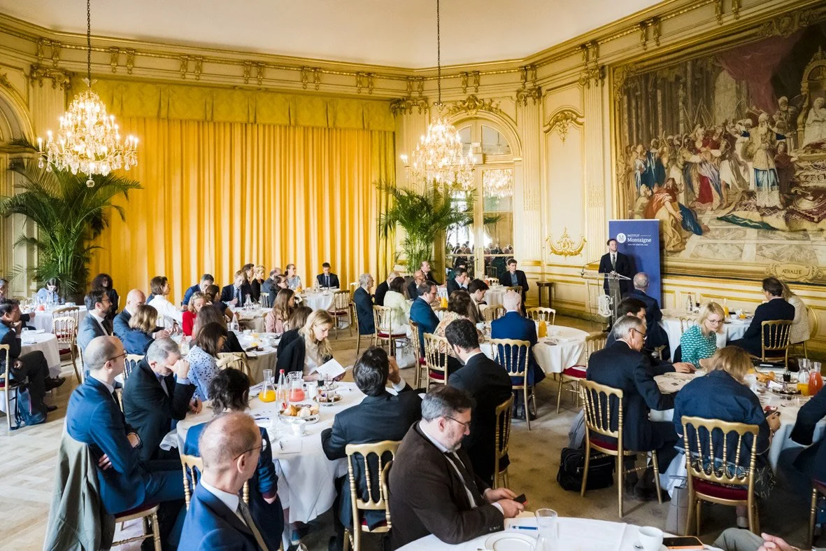Une salle de conférence élégante avec des participants assis à des tables rondes, un homme parle à un podium, décorations luxueuses avec lustres, grands miroirs et une peinture sur le mur.