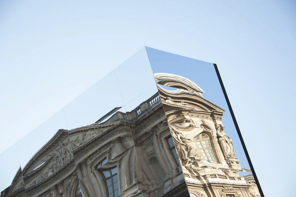 Reflet d'un bâtiment historique avec des sculptures, dans une surface miroir, sous un ciel clair.