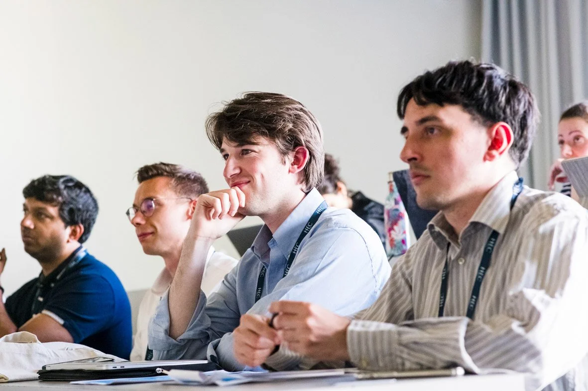 Jeunes adultes attentifs lors d'une conférence, assis à une table avec des documents et portants des badges d'identification.