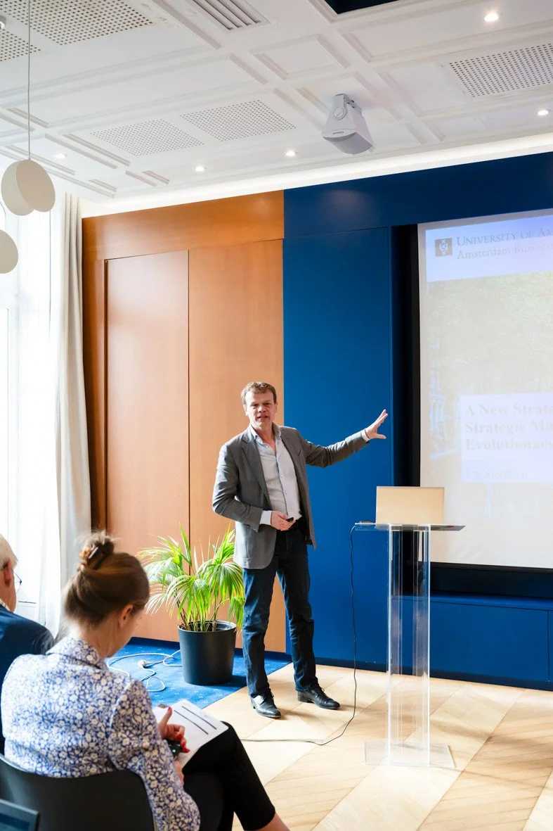 Un homme en costume présentant lors d'une conférence dans une salle moderne avec un écran et quelques personnes assises.