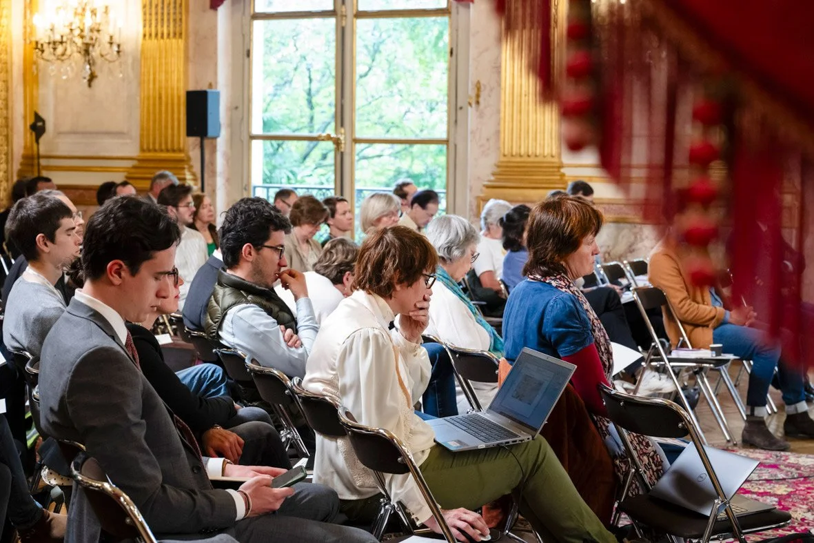 Audience lors d'une conférence dans une salle ornée de dorures et de grands rideaux rouges, avec des personnes concentrées, certaines utilisant un ordinateur portable ou un téléphone.