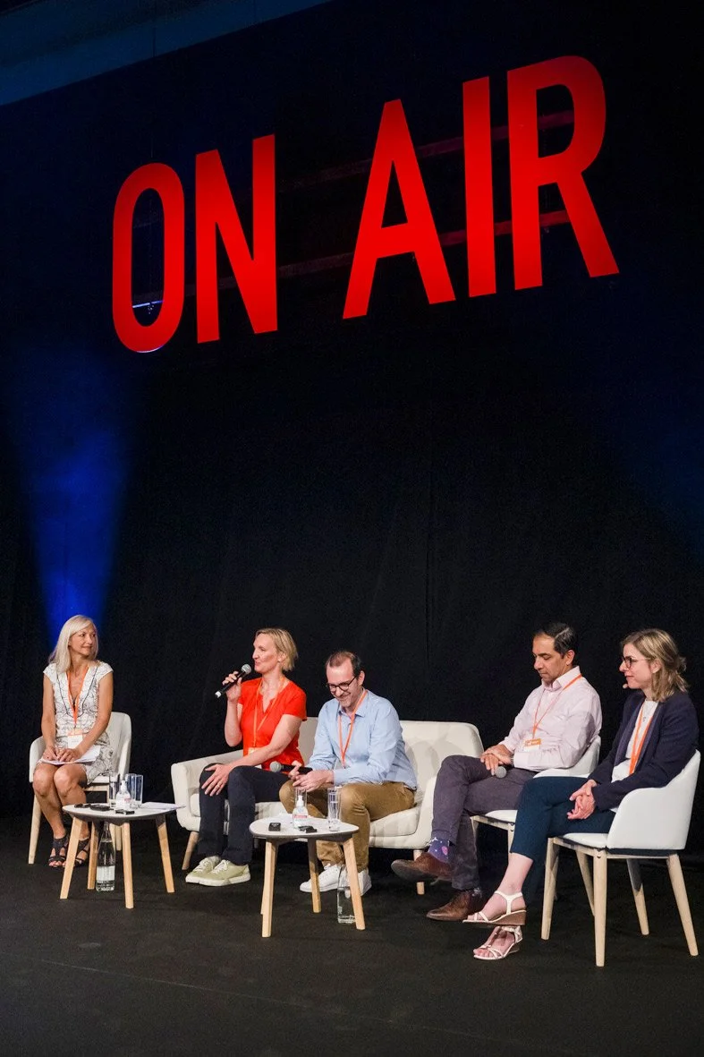 Cinq personnes assises sur un podium lors d'une discussion, avec un grand panneau rouge lisant 'ON AIR' en arrière-plan.