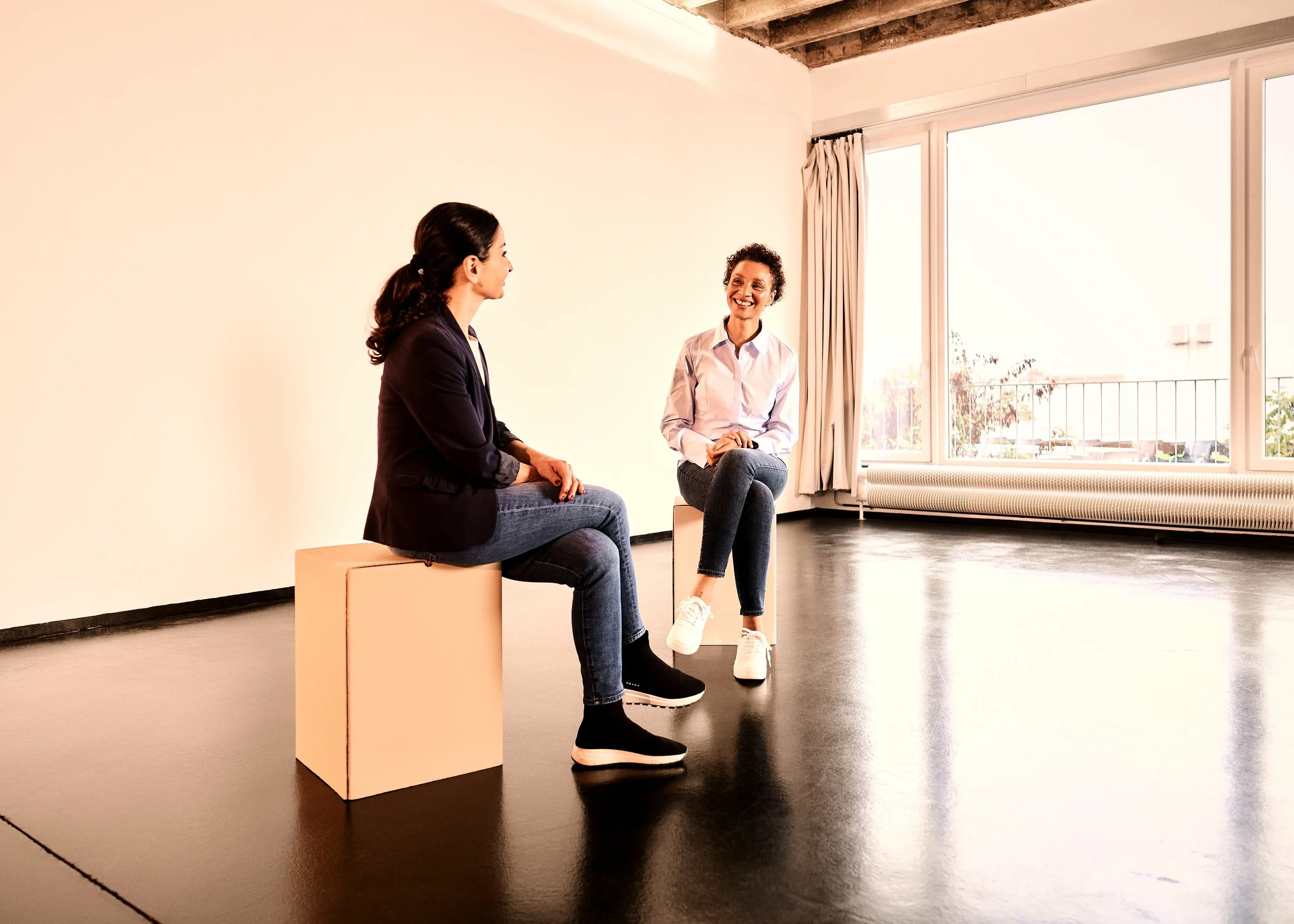 Gaëlle Georges und Teilnehmende beim Resilienz-Coaching. Sie sitzen in einem hellen Raum auf Blockhockern und führen ein Gespräch. Eine Frau trägt eine dunkle Jacke und eine andere ein weißes Hemd, beide lachen.