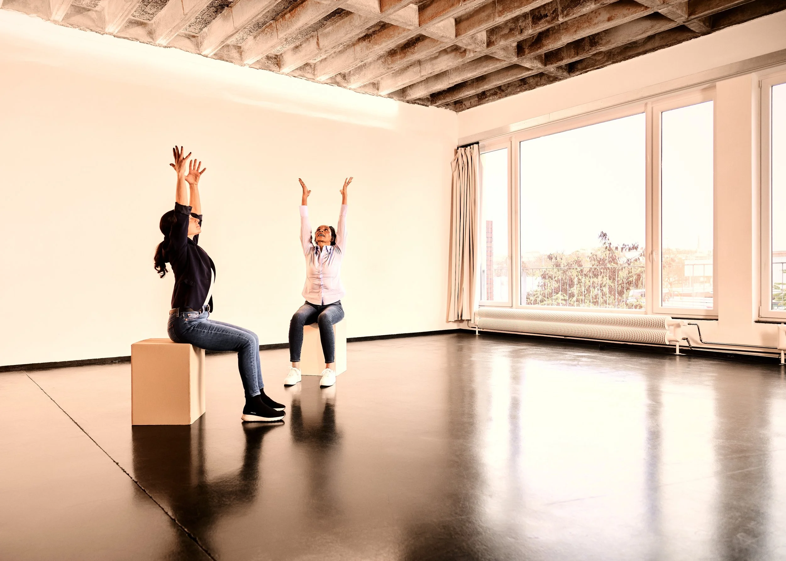 Zwei Frauen sitzen auf Sitzwürfeln in einem leeren Raum mit großen Fenstern, sie üben Yoga oder andere Dehnübungen, die Sonne scheint hinein.