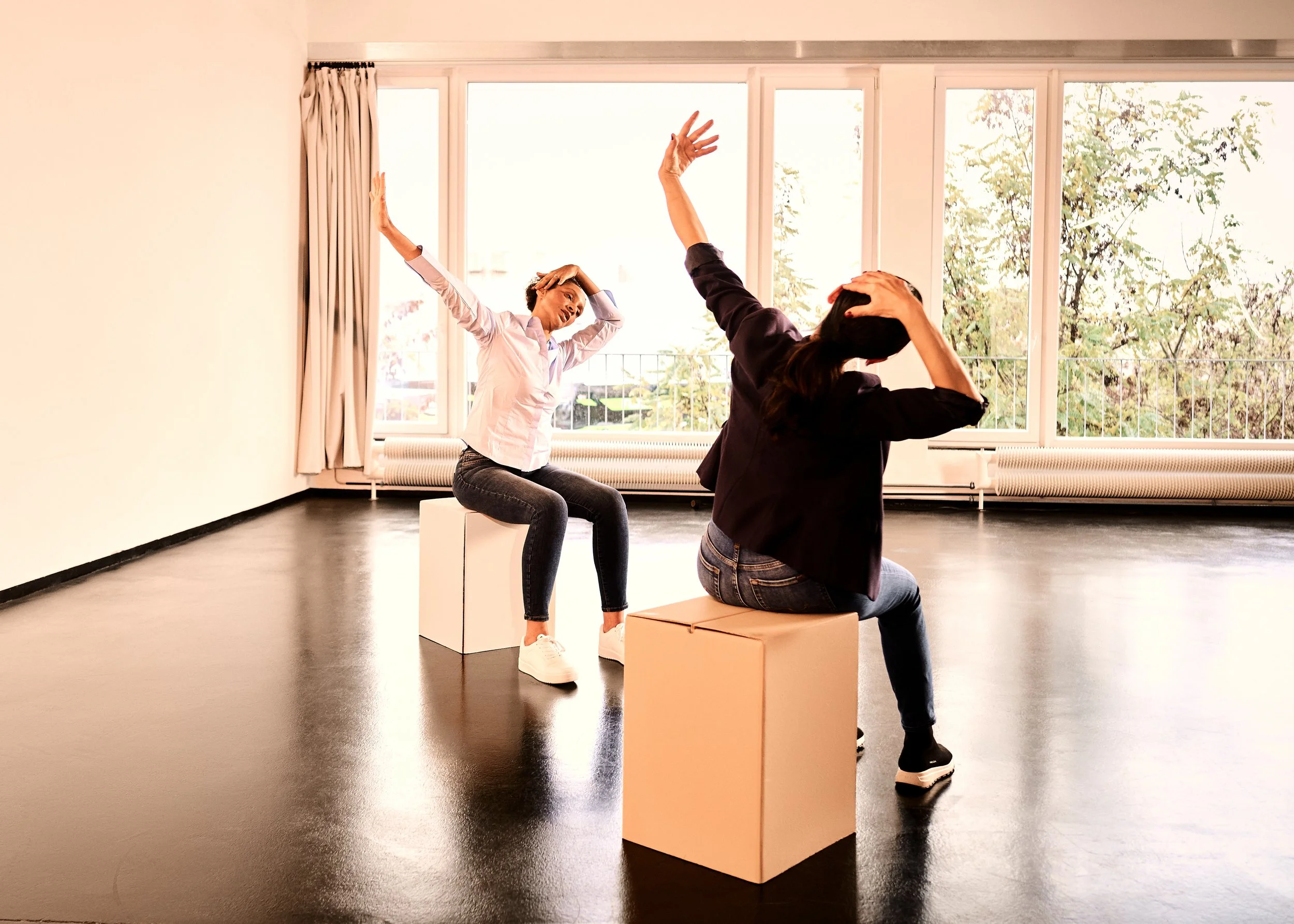 Gaëlle Georges sitzt mit einer Klientin auf weißen Quader in einem hellen Raum. Sie entspannen , während sie nach oben strecken und den Kopf in den Händen halten.