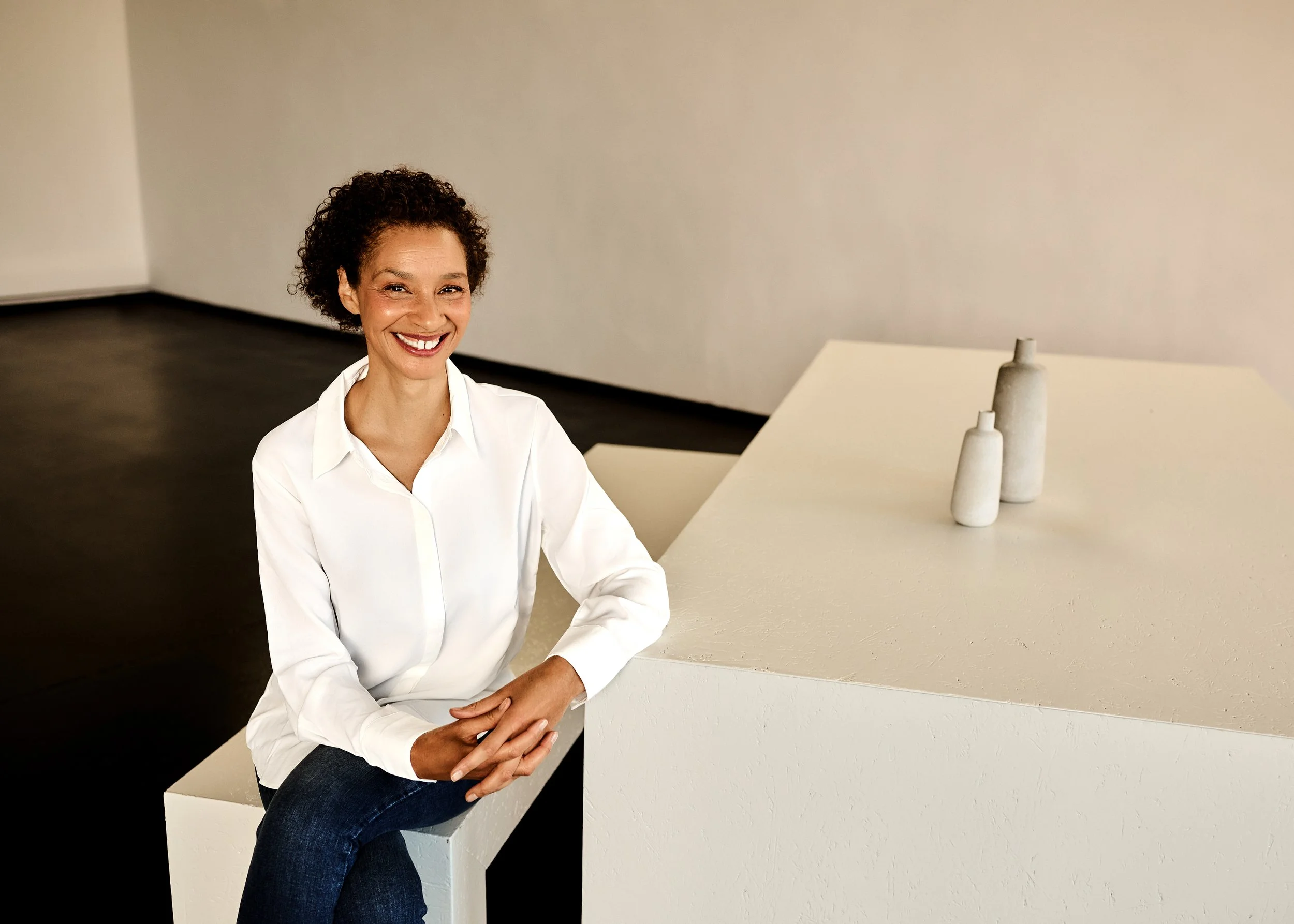 Eine lächelnde Frau mit braunen, lockigen Haaren sitzt auf einem weißen Podest in einem minimalistischen Raum mit weißen Wänden, neben einem weißen Tisch mit drei stilisierten Vase-Dekorationen.