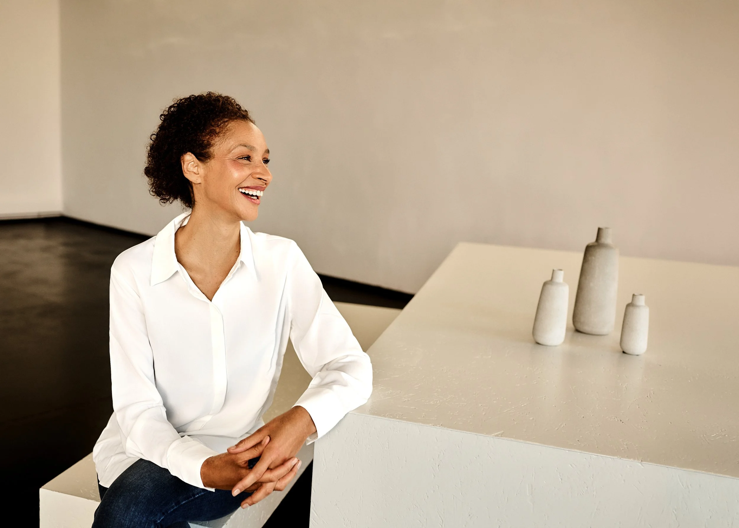 Eine lachende Frau sitzt in einem Museum neben einer weißen Skulptur mit mehreren kleinen Vasen.