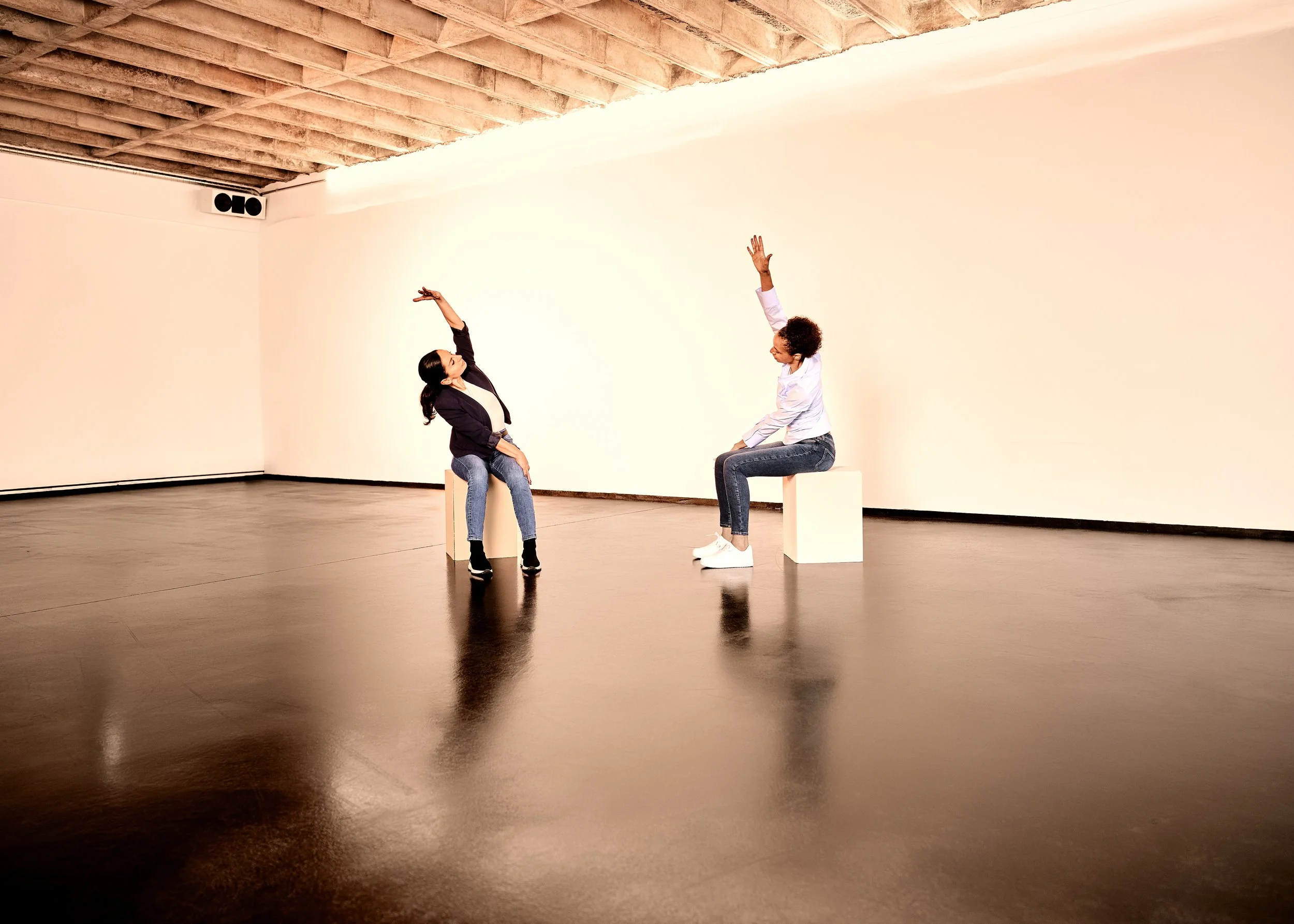 Zwei Frauen sitzen auf weißen Quadern in einem großen, leeren Raum mit Holzdecke, die eine scheint eine Frage zu stellen, die andere antwortet.