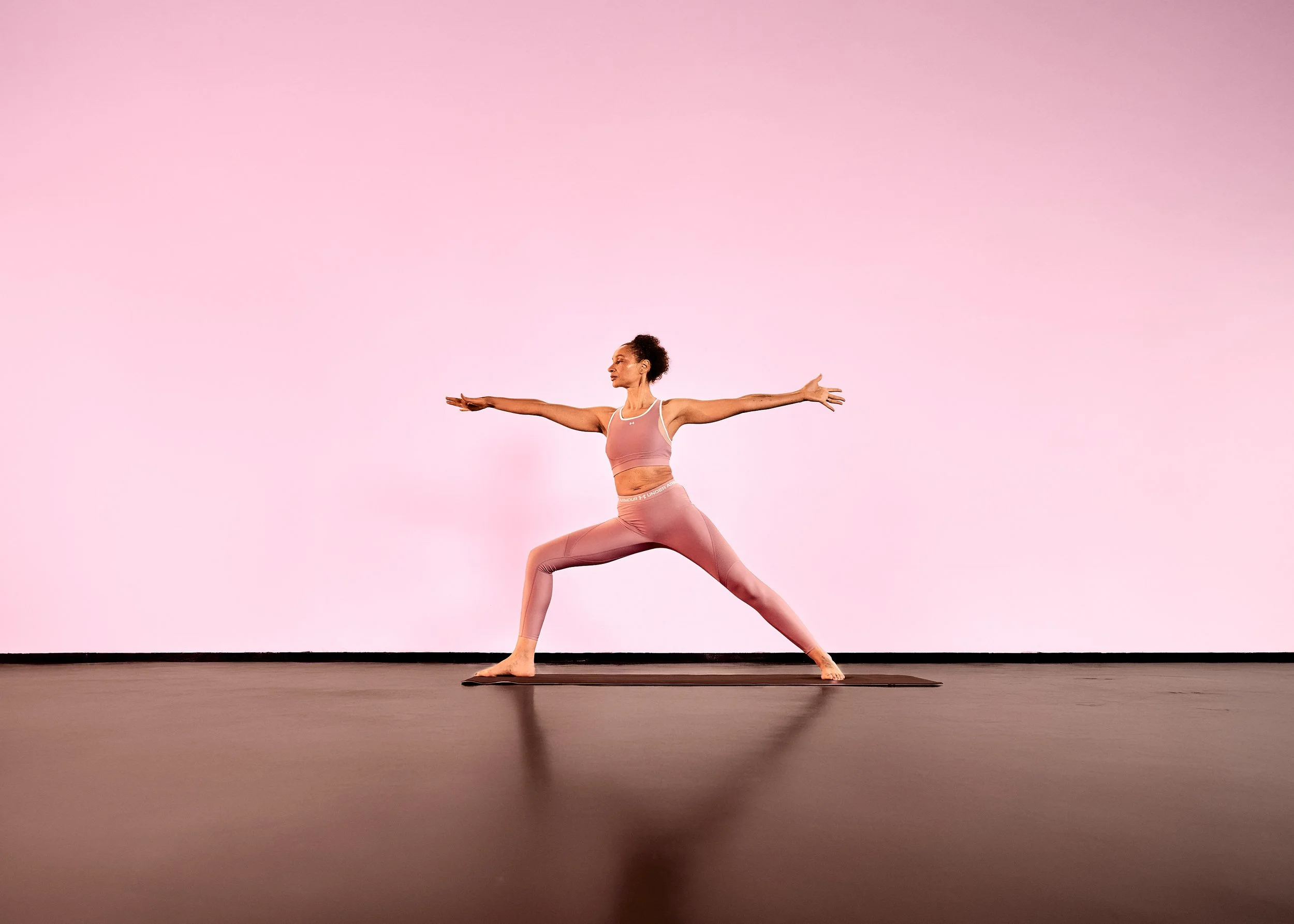 Gaëlle Georges macht Yoga in einer Pose auf einer Yogamatte vor einem pinken Hintergrund.