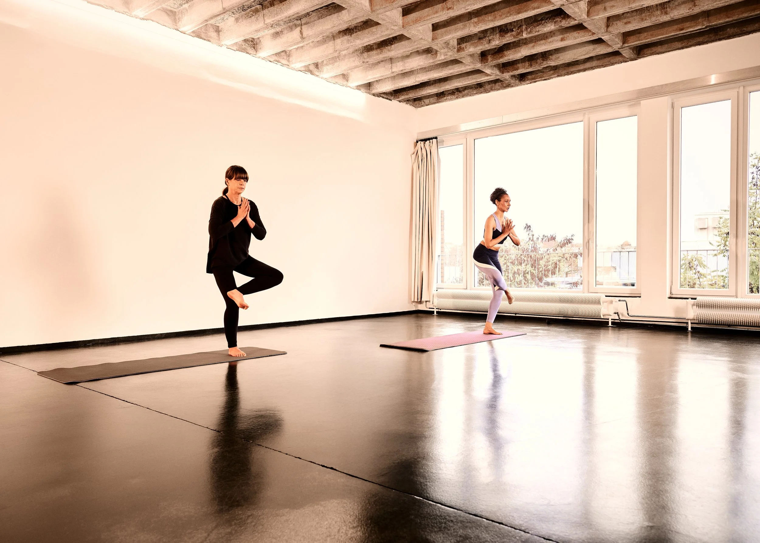 Zwei Frauen machen Yoga in einem hellen Raum mit großen Fenstern und Holzbalkendecke. Beide stehen im Baum-Haltung auf Yogamatten, mit gefalteten Händen vor der Brust.
