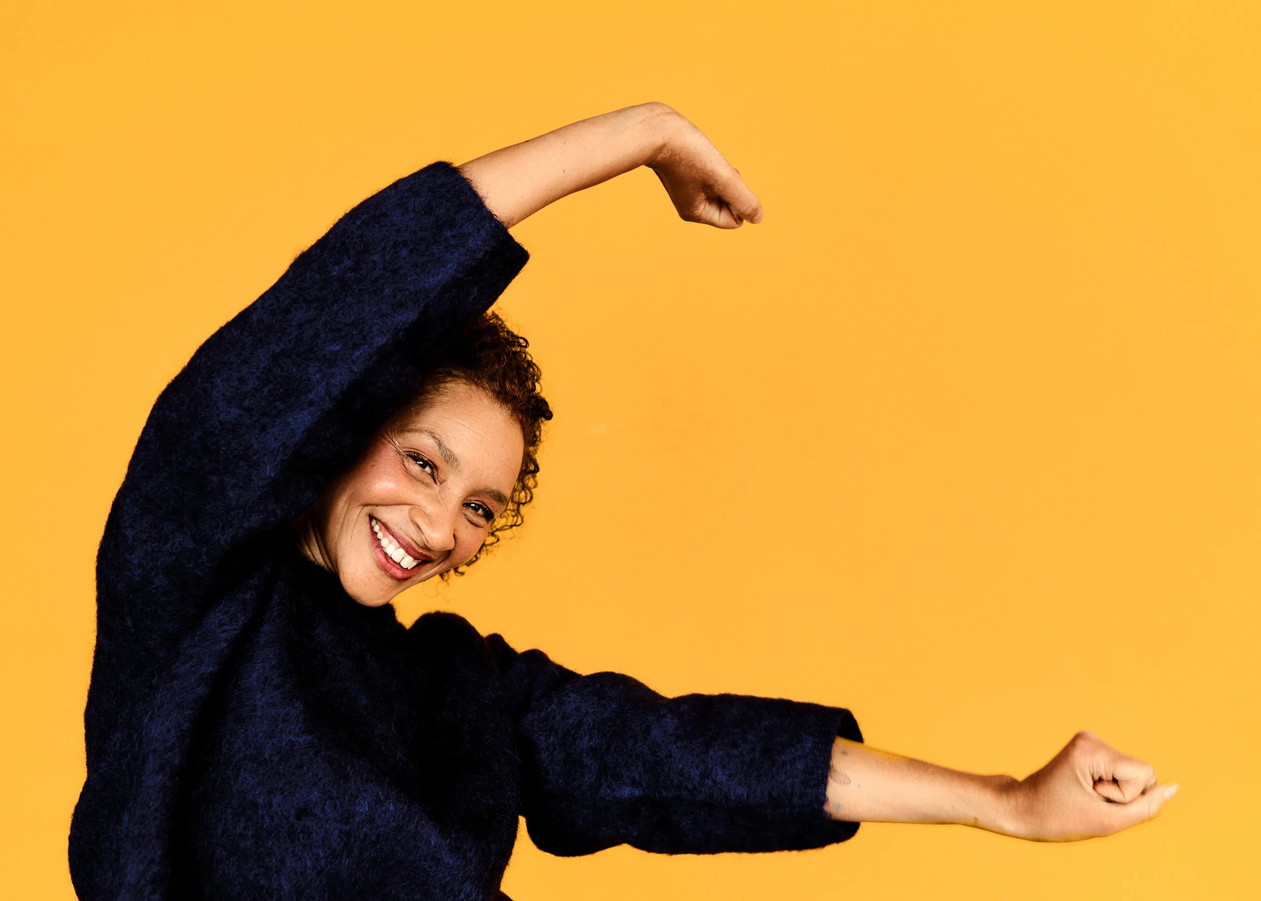 Gaëlle Georges in dunklem Pullover vor einem gelben Hintergrund und die Arme nach oben streckt.