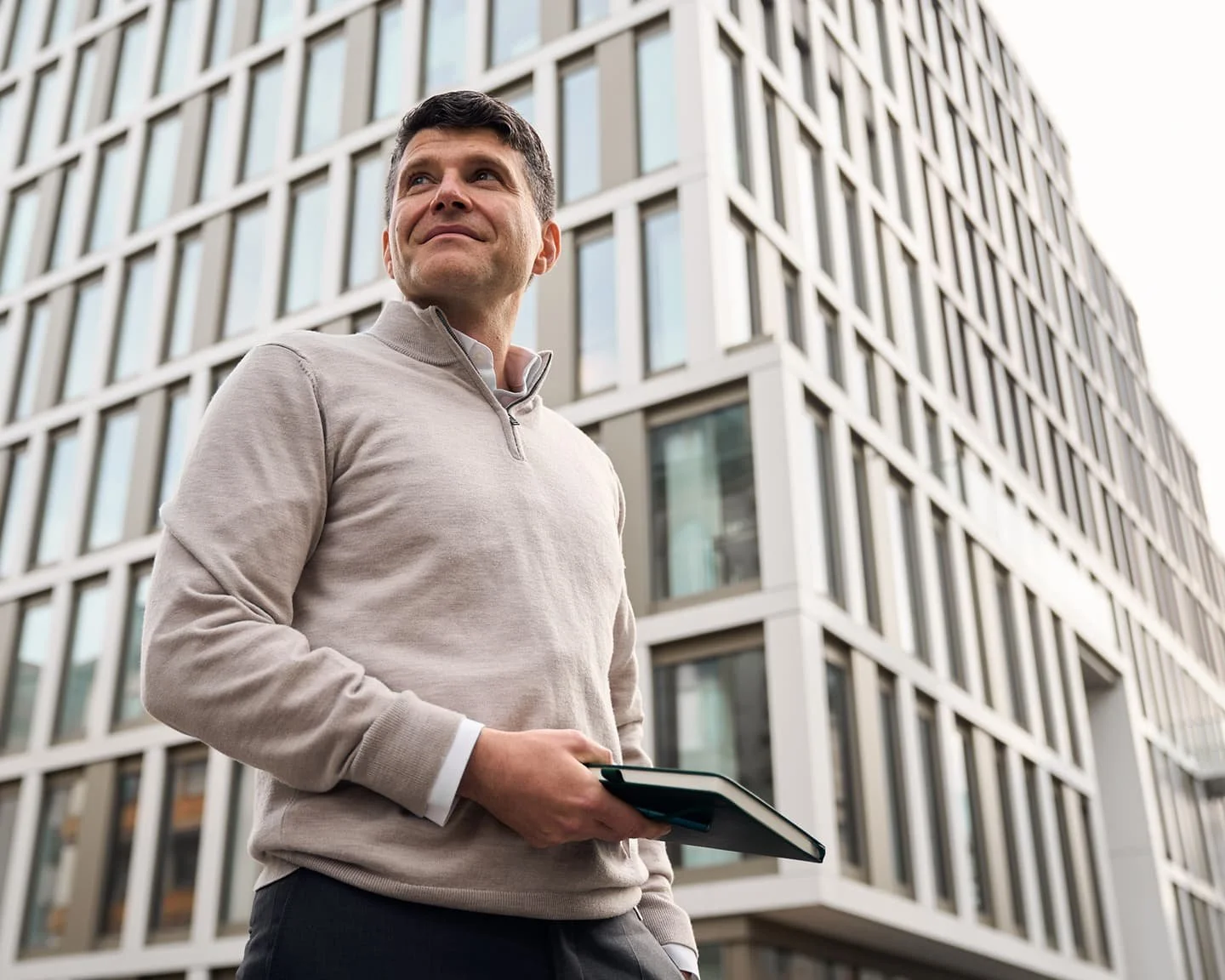 Stephen von der Brüggen steht vor einem modernen Bürogebäude, trägt einen hellen Pullover und hält ein Buch oder einen Ordner in der Hand.