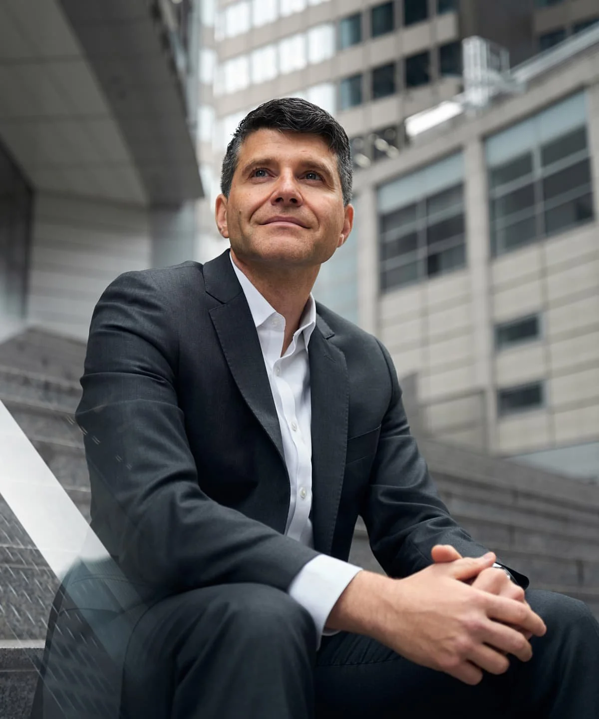 Stephen von der Brüggen in einem dunklen Anzug sitzt auf einer Treppe vor einem modernen Bürogebäude, die Stadt Frankfurt im Hintergrund.