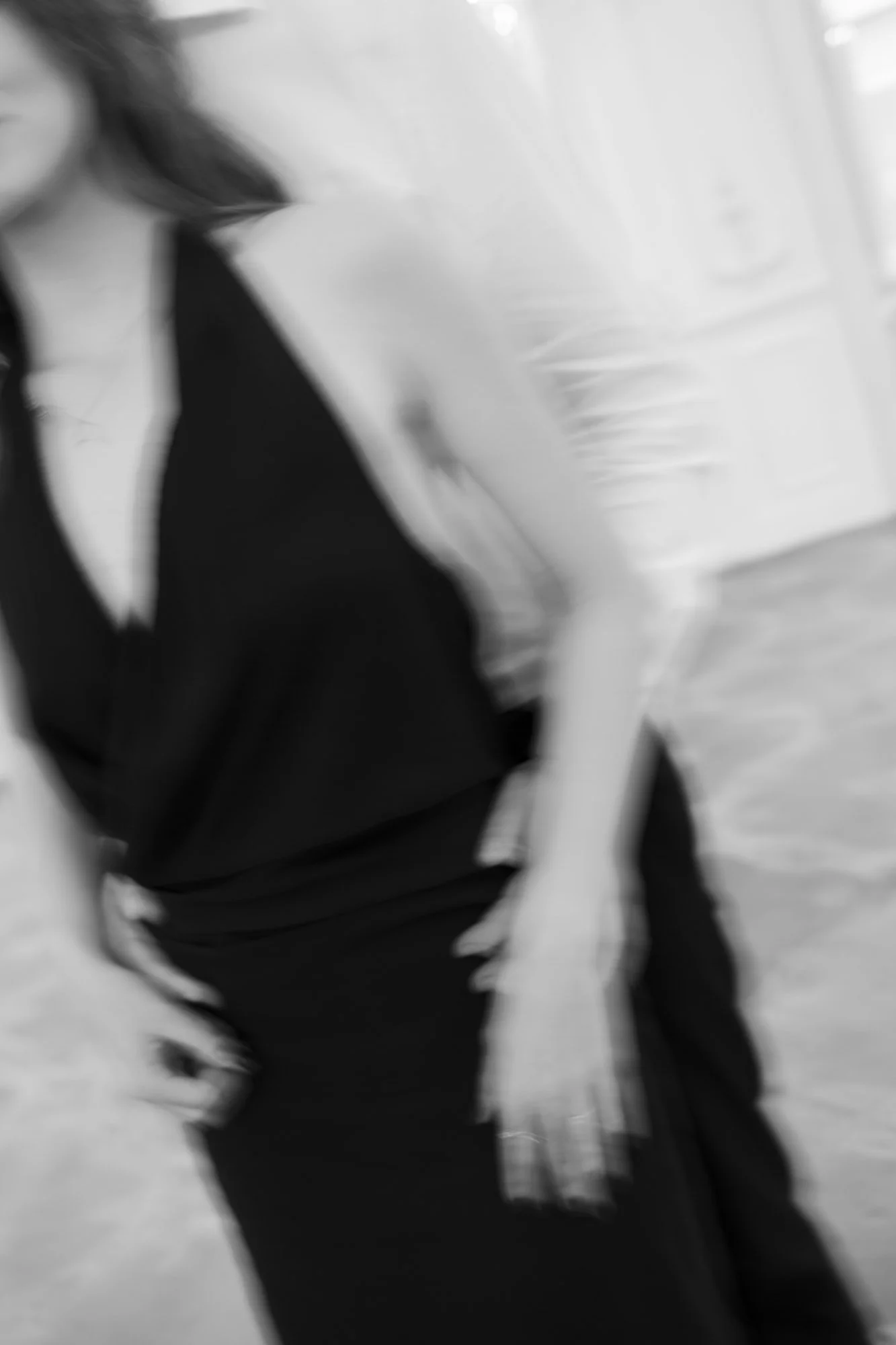 A person wearing a black shirt and black pants, with part of their arm visible, standing in an indoor space with a white wall and a window in the background.