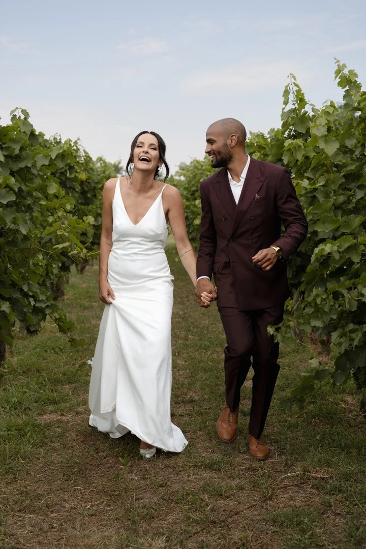 A couple in wedding attire walking through a vineyard, holding hands and smiling.
