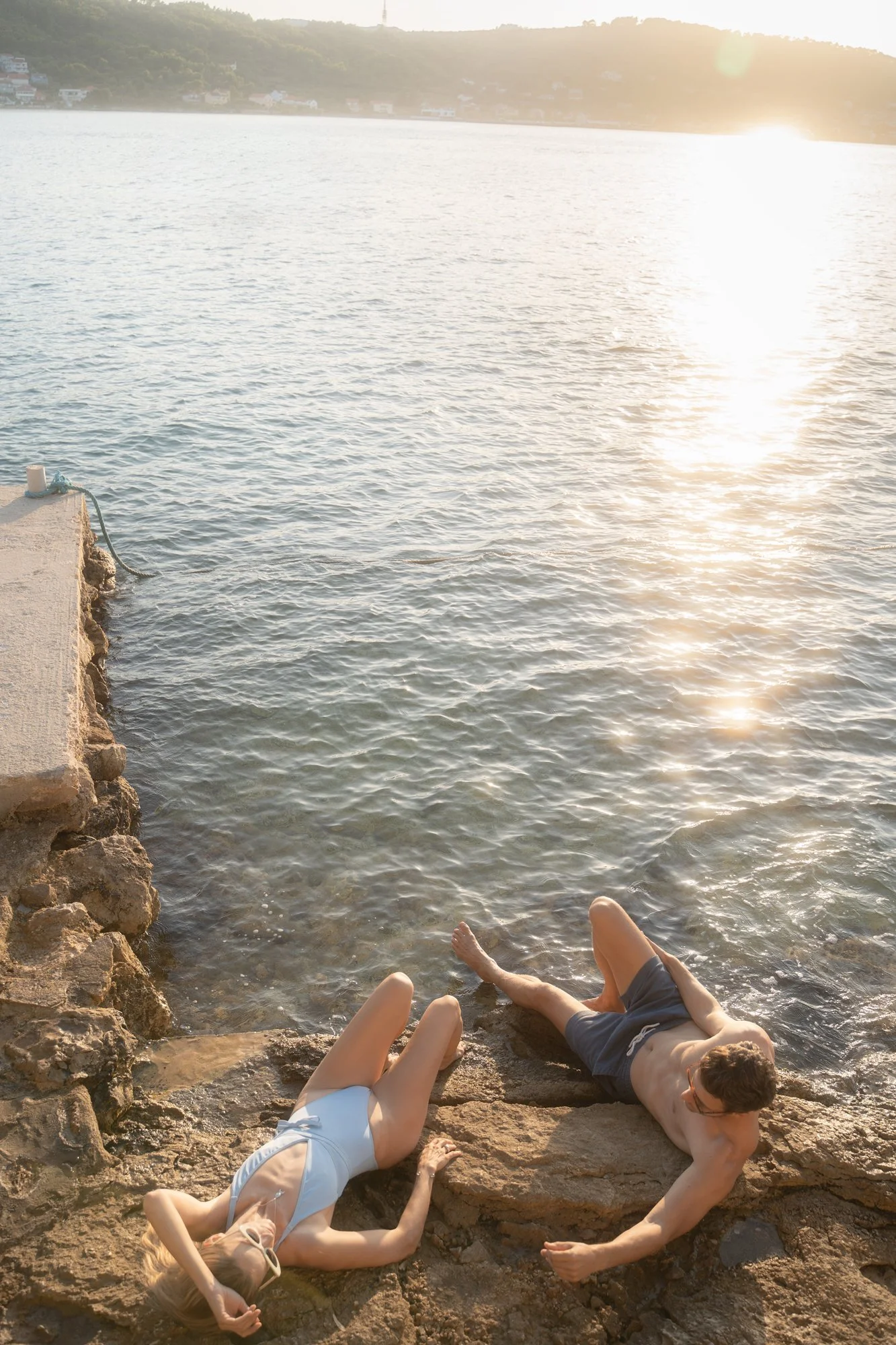 A man and a woman relaxing on rocks by the water's edge during sunset, with the sun reflecting on the water and distant hills in the background.