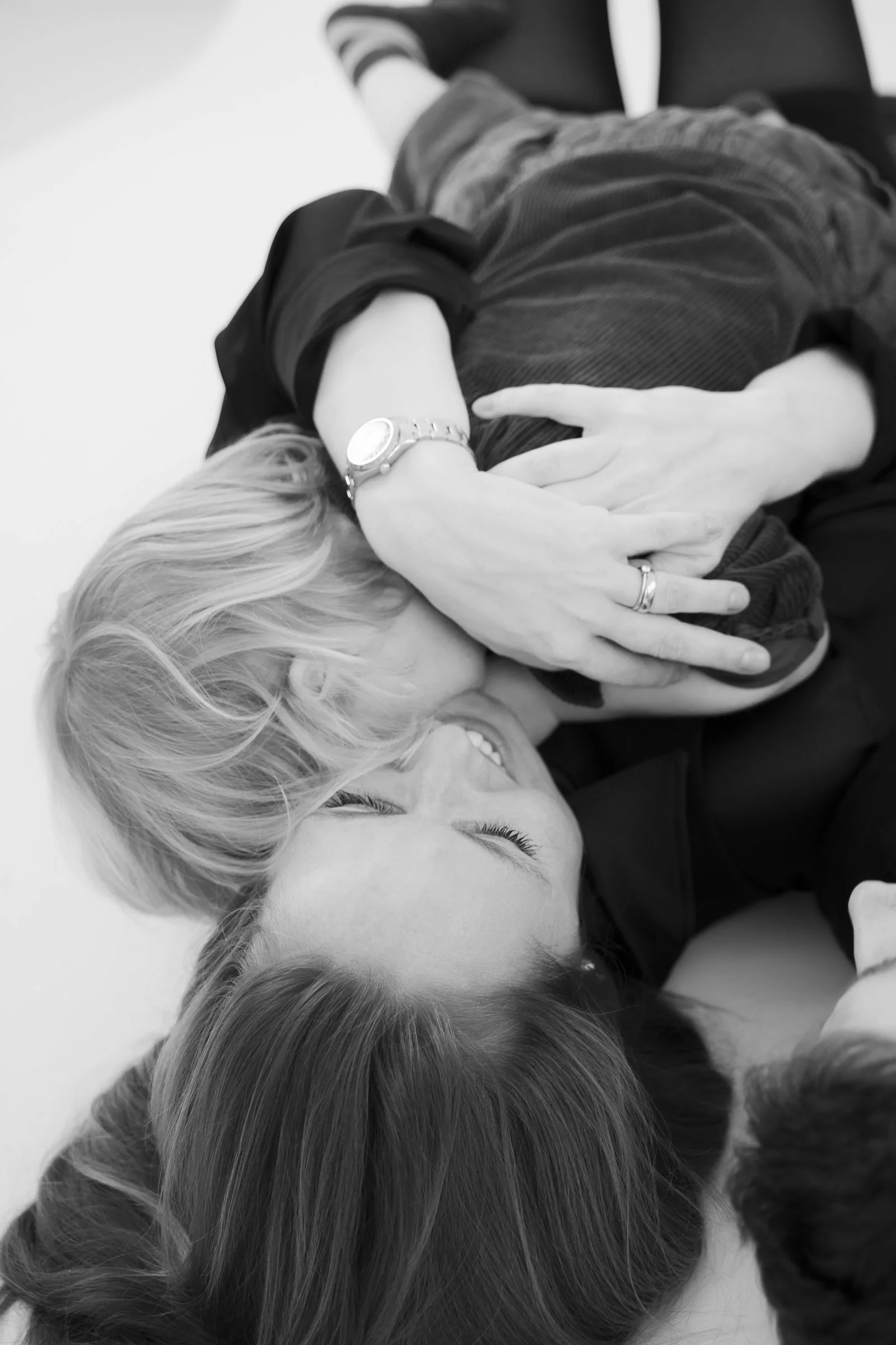 Two women lying on their sides, laughing and hugging each other, in a playful, intimate pose.
