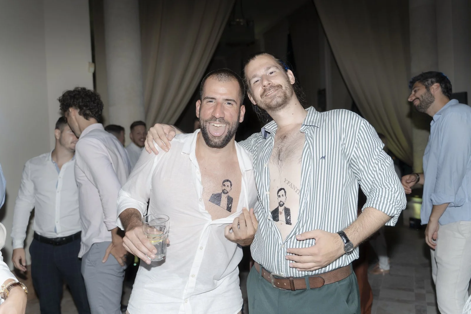 Two men at a party, wearing white and striped shirts, showing tattoos of a man's portrait on their chests, smiling and posing for the camera.