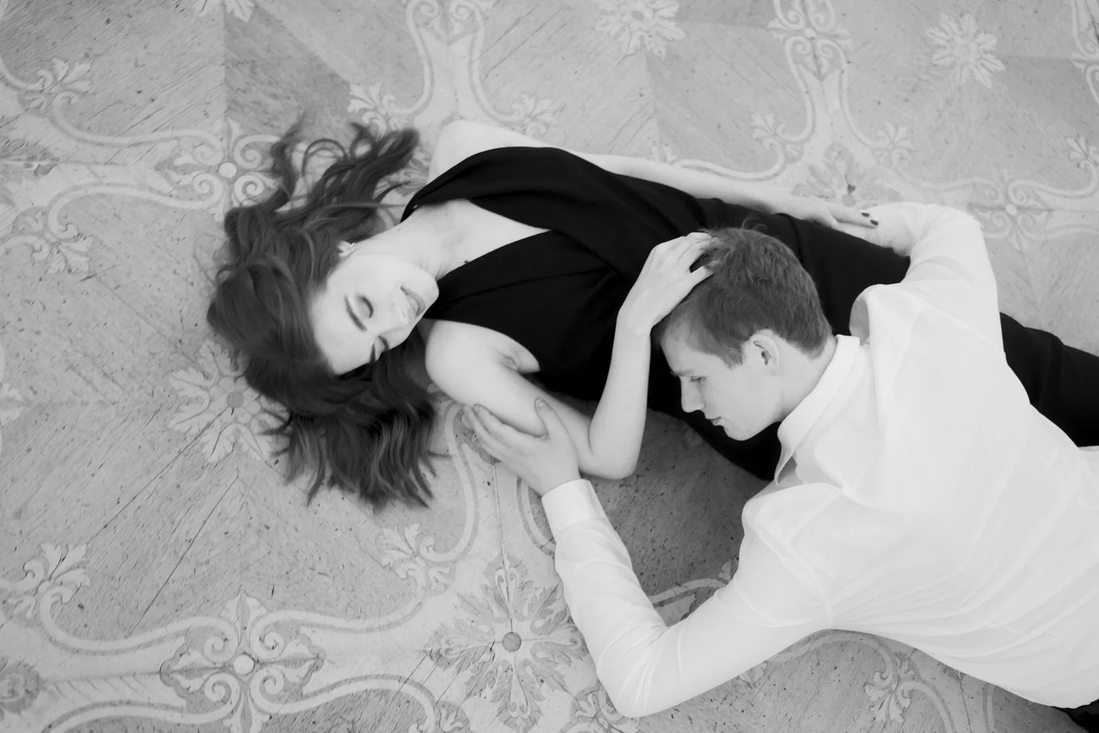 A man and woman lying on the floor, the woman appears to be unconscious or resting, while the man is holding her arm and touching her face gently.