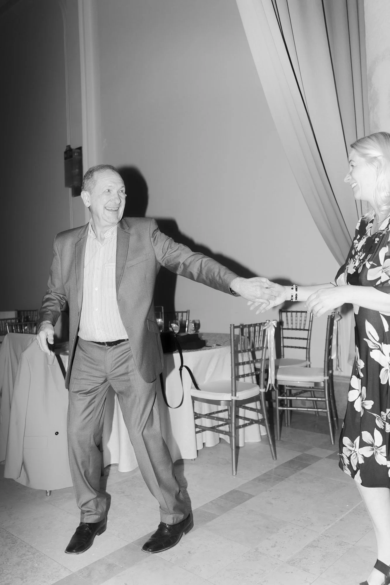 An elderly man in a suit with a woman in a floral dress sharing a joyful dance or moment together in a decorated event space.