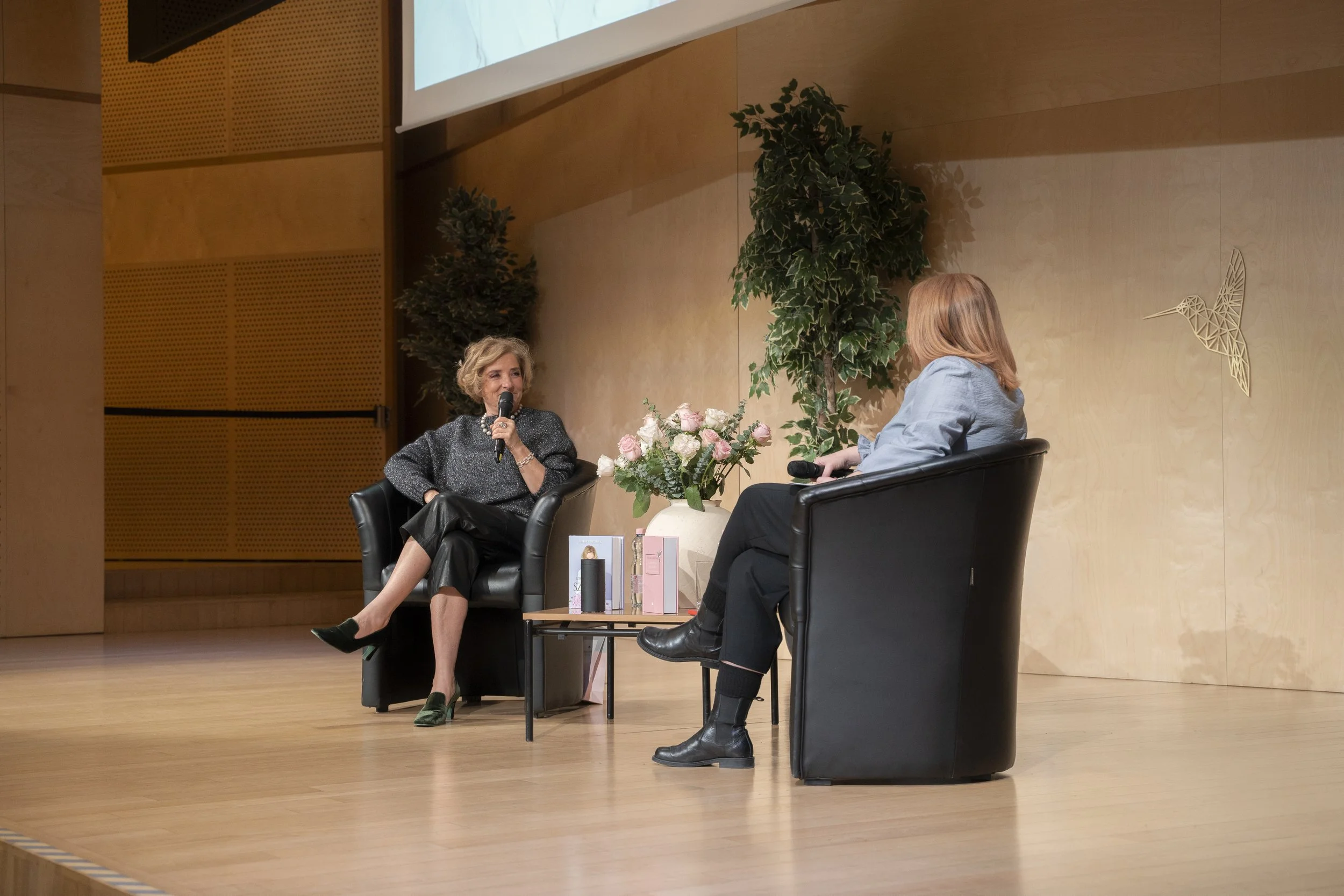 Two women having a conversation on a stage, sitting in black armchairs with a small table between them holding pink and white flowers in a vase and books, with large potted plants behind them and a hummingbird wall decoration on the wooden wall.