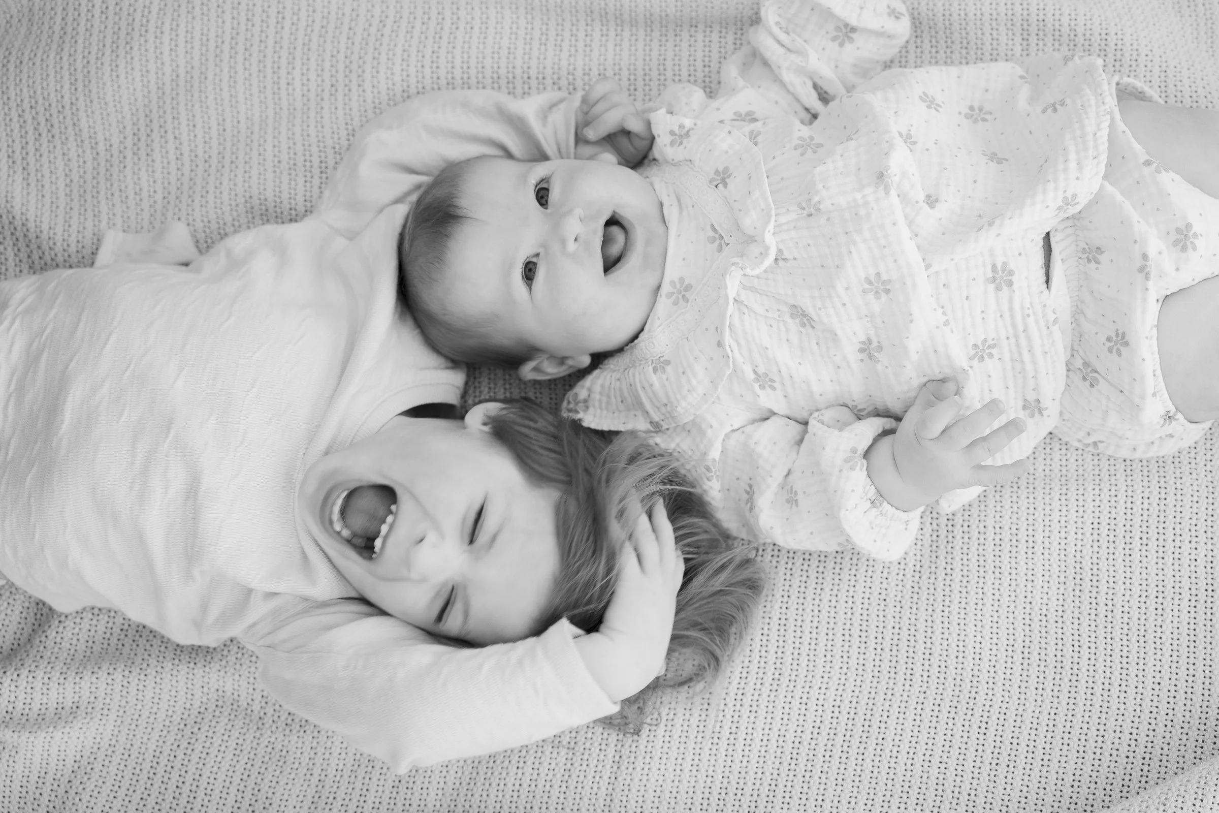 Black and white photo of two children lying on a textured bed, one boy and one girl, smiling and laughing.