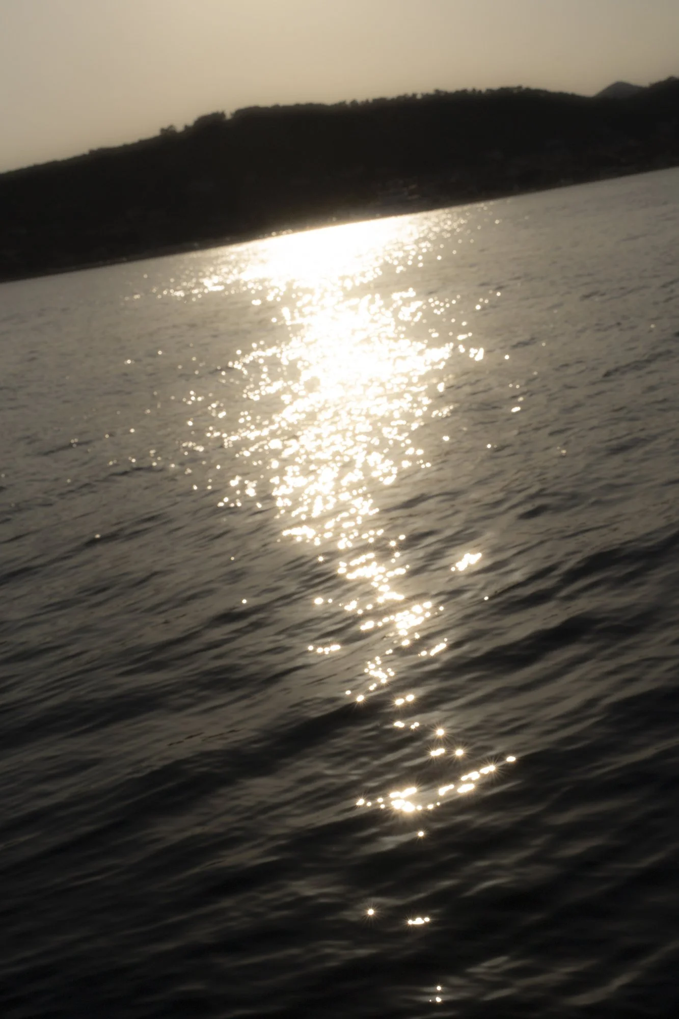 Sunset over a body of water with ripples, reflecting sunlight, with a distant hill or mountain on the horizon.