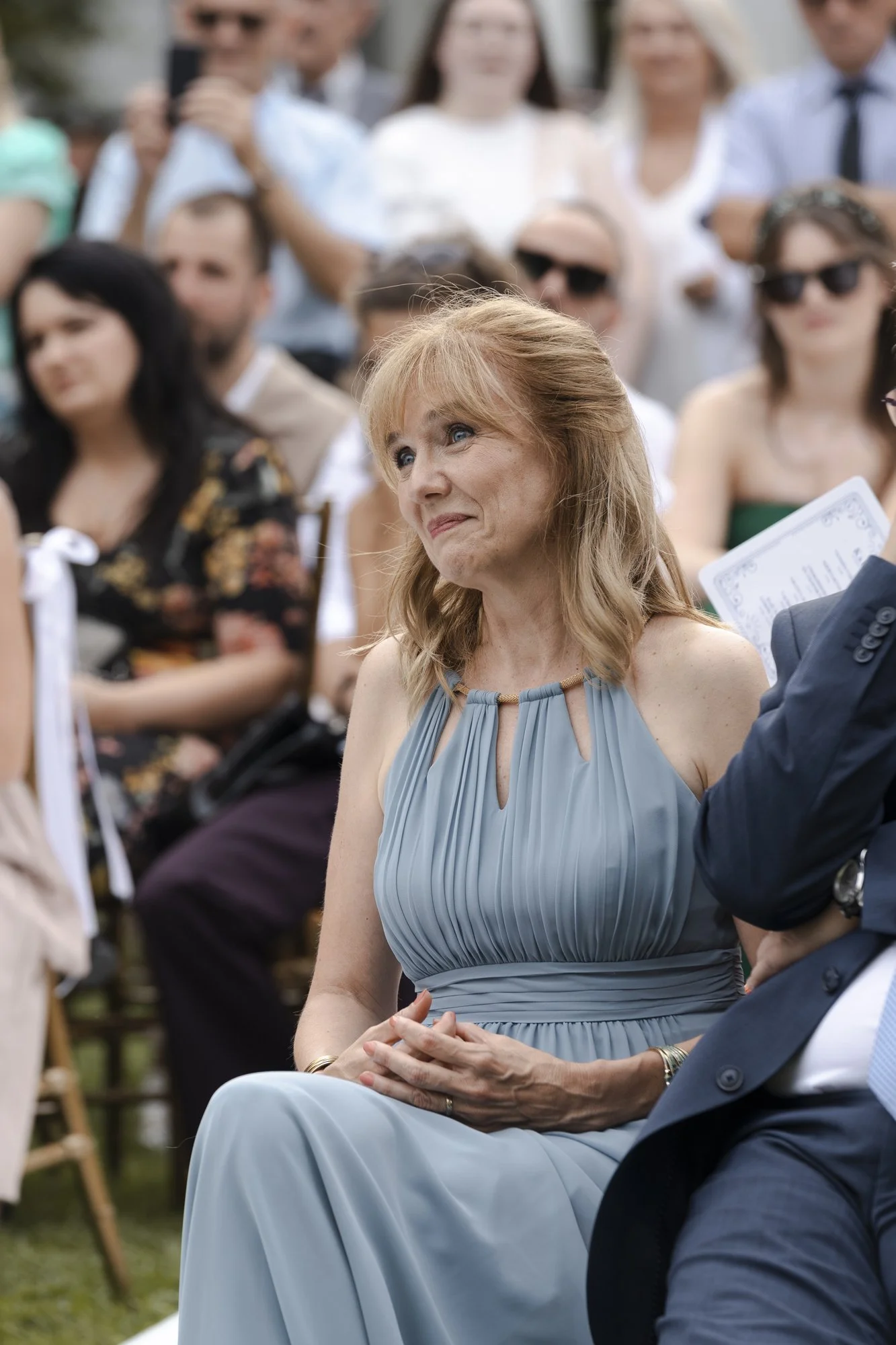 A middle-aged woman with red hair and blue eyes, wearing a light blue pleated dress, is sitting outdoors among a group of people. She appears to be emotional, possibly crying, and is looking towards someone or something in front of her.