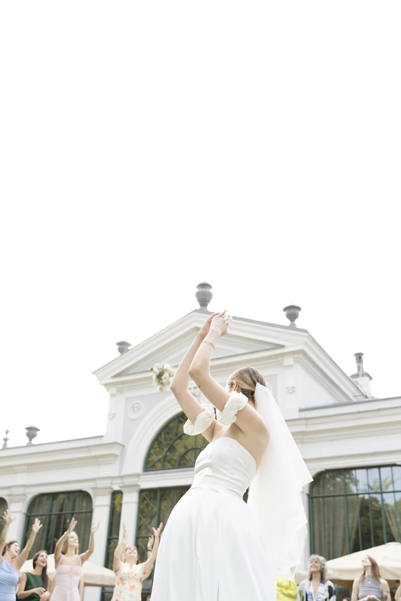 A bride in a white wedding dress and veil dances outdoors among a group of women, in front of a white building with arched windows