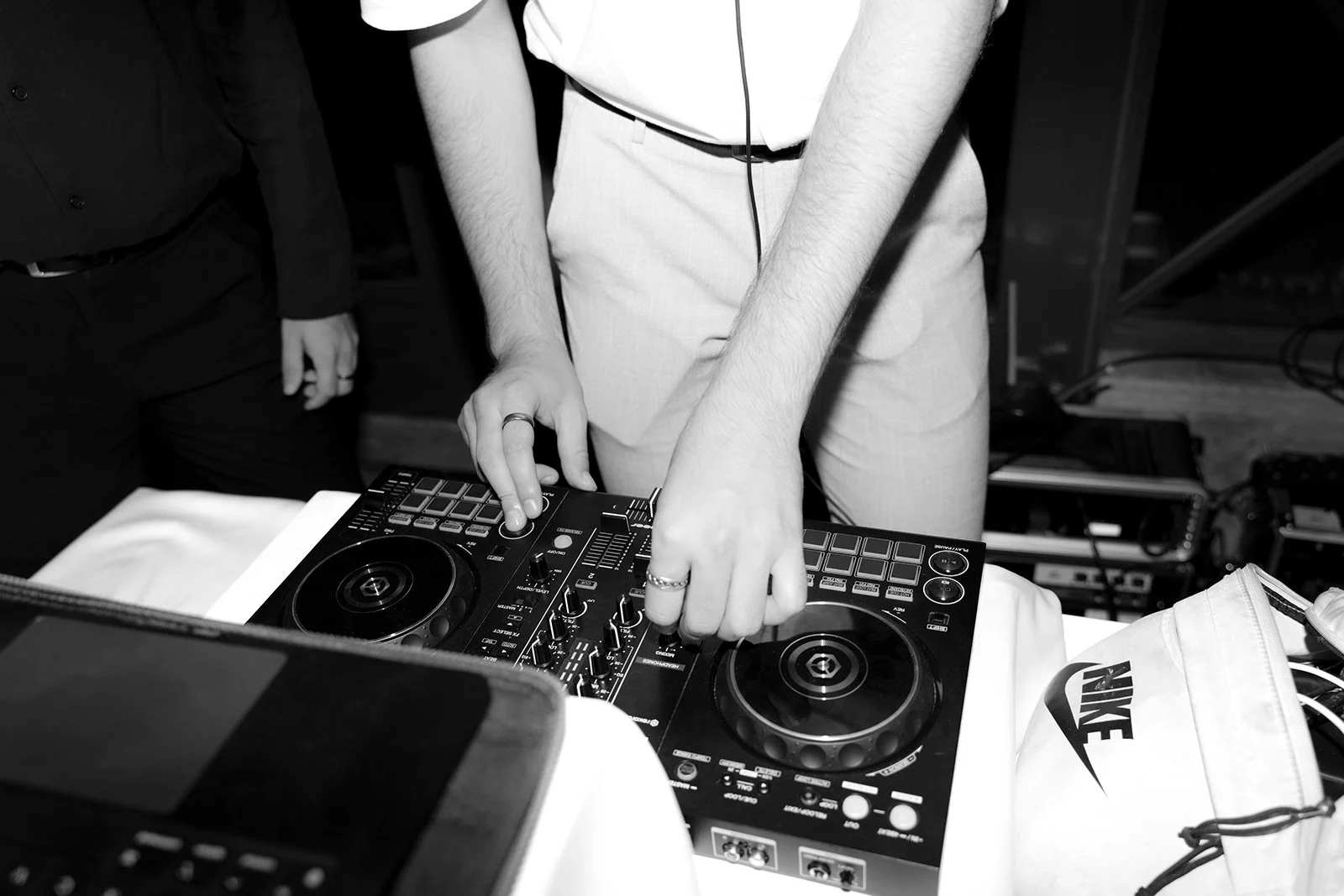 Person using DJ equipment with two turntables and a mixer, with another person standing nearby, in a black and white photo.