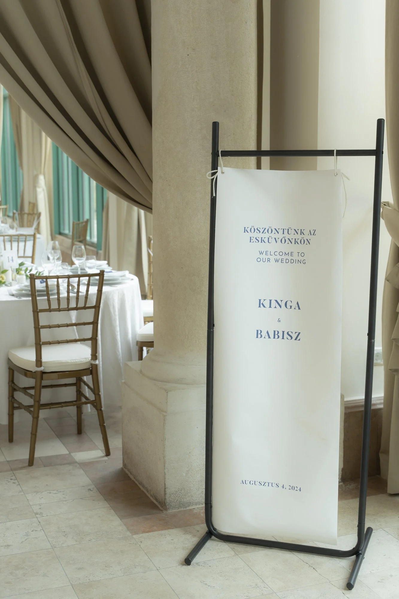 Wedding welcome sign with Hungarian and English text, displaying the names Kinga and Babisz, dated August 4, 2024, in an elegant indoor setting with tables and chairs.