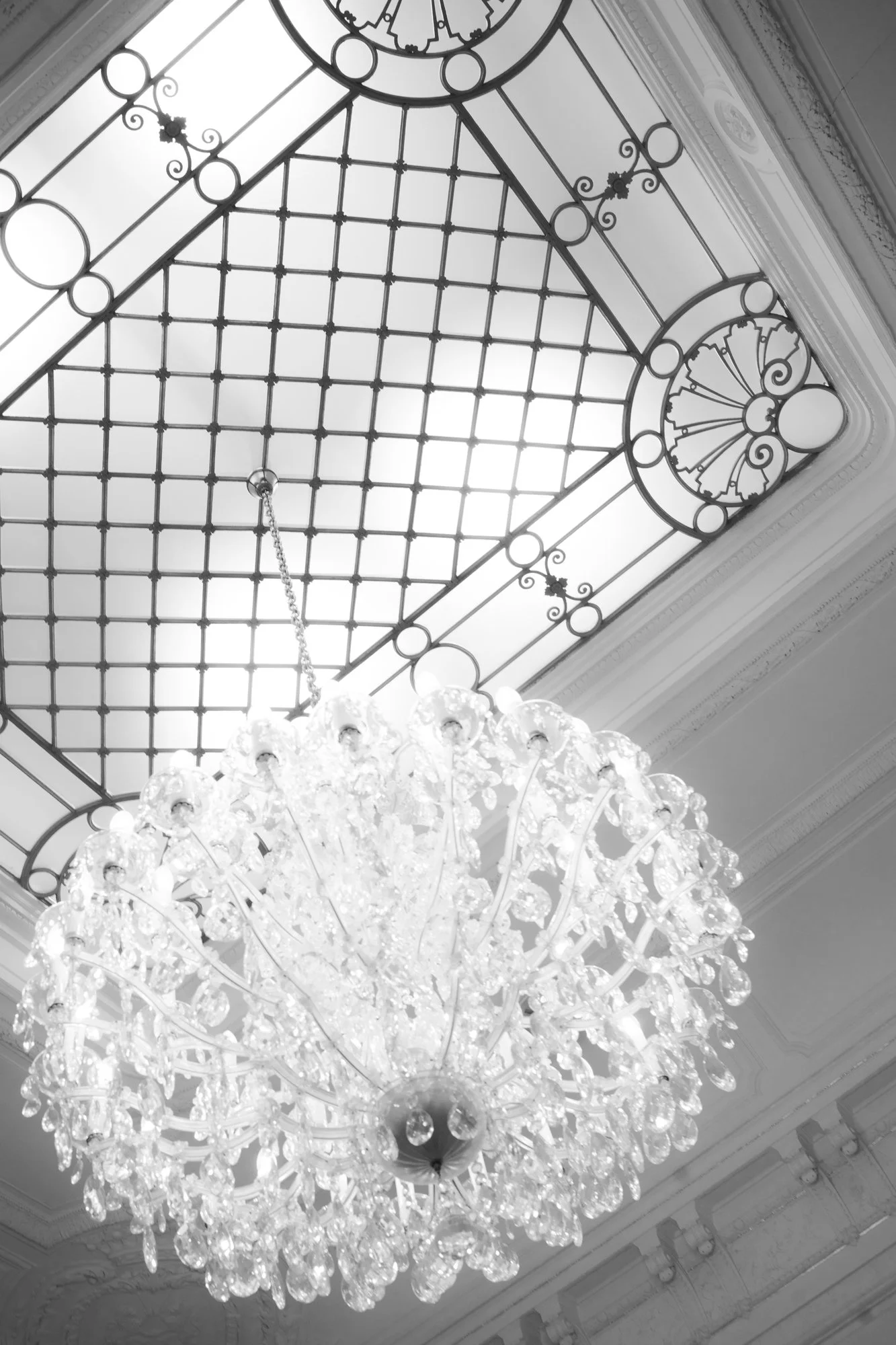 A crystal chandelier hanging from an ornate ceiling with a large decorative glass skylight.