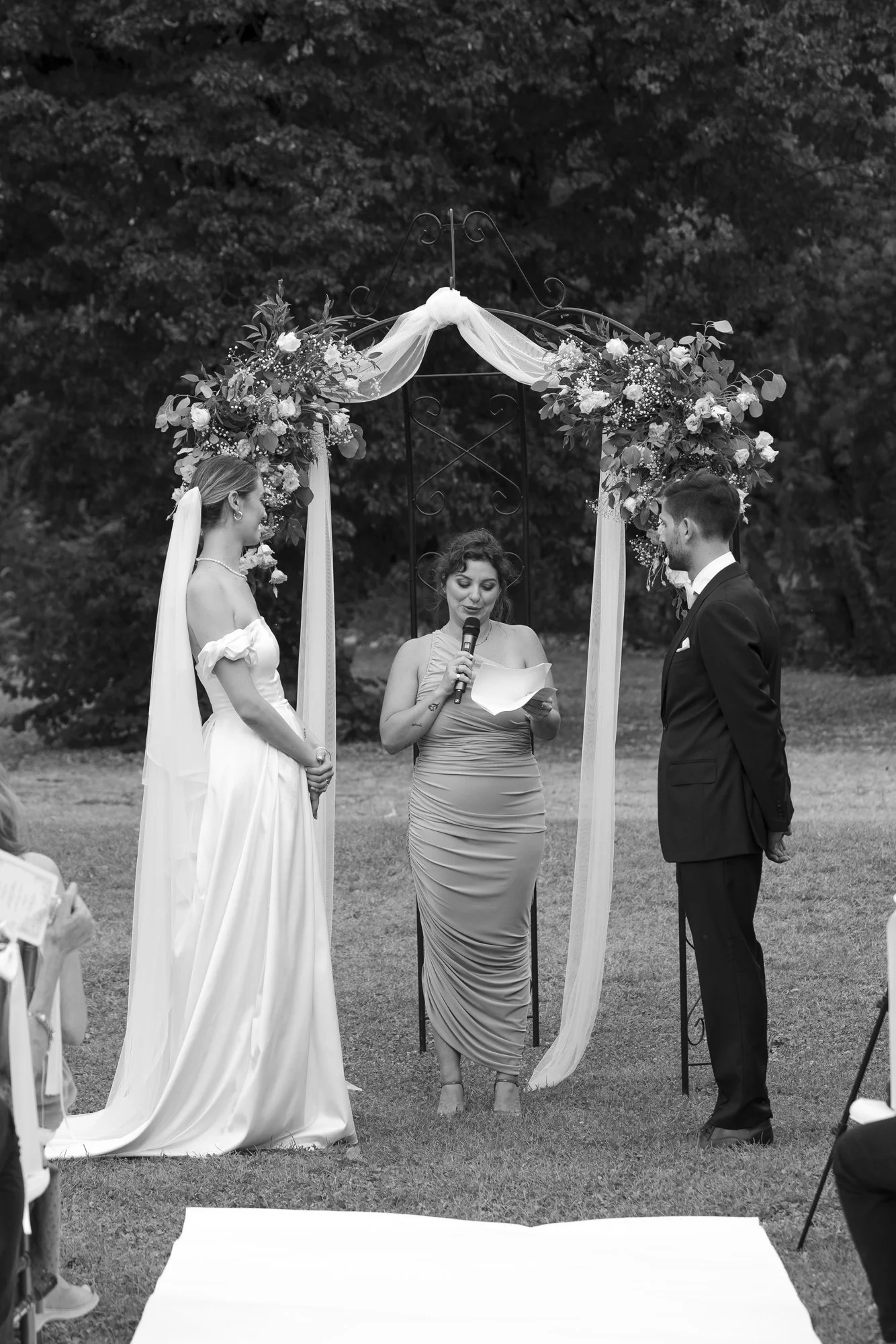 A couple getting married outdoors during the daytime, standing in front of an arch decorated with flowers and draped fabric, with a woman officiating the ceremony reading from a paper while holding a microphone.